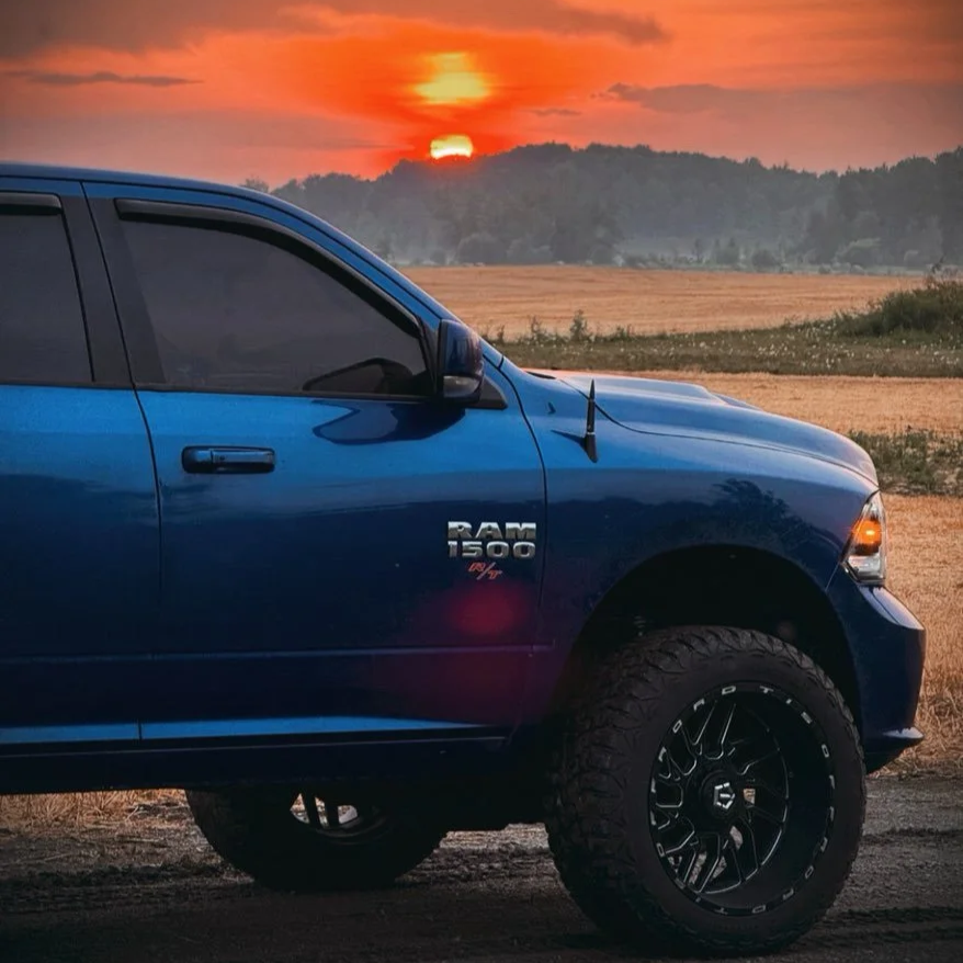 A blue Ram 1500 pickup truck parked on a dirt road during sunset, with a sunset sky and distant hills in the background.