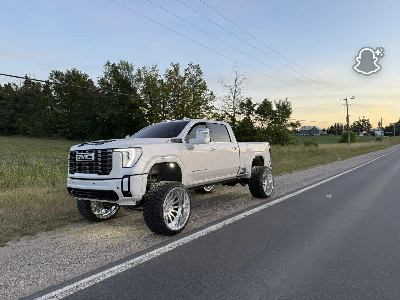 A white GMC truck with oversized wheels parked on the side of a rural road during dusk, with trees and houses in the background.