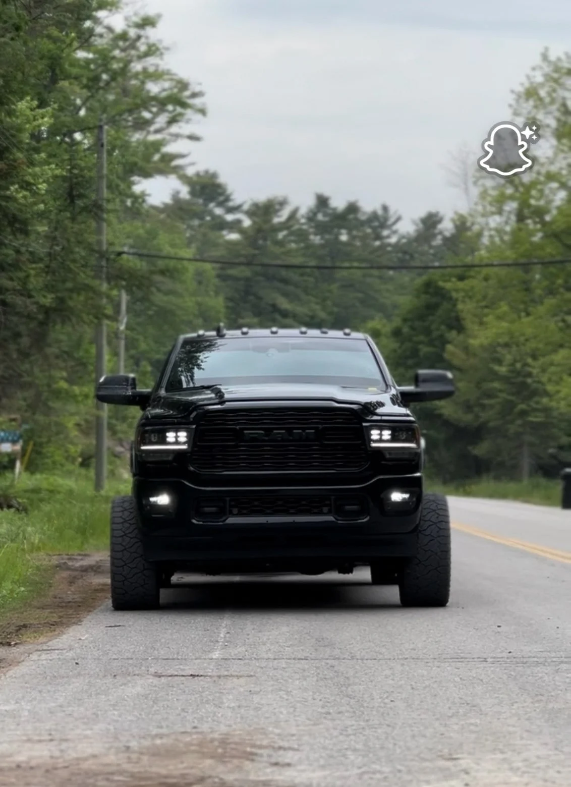 Black RAM truck parked on a rural road with green trees and utility poles in the background.