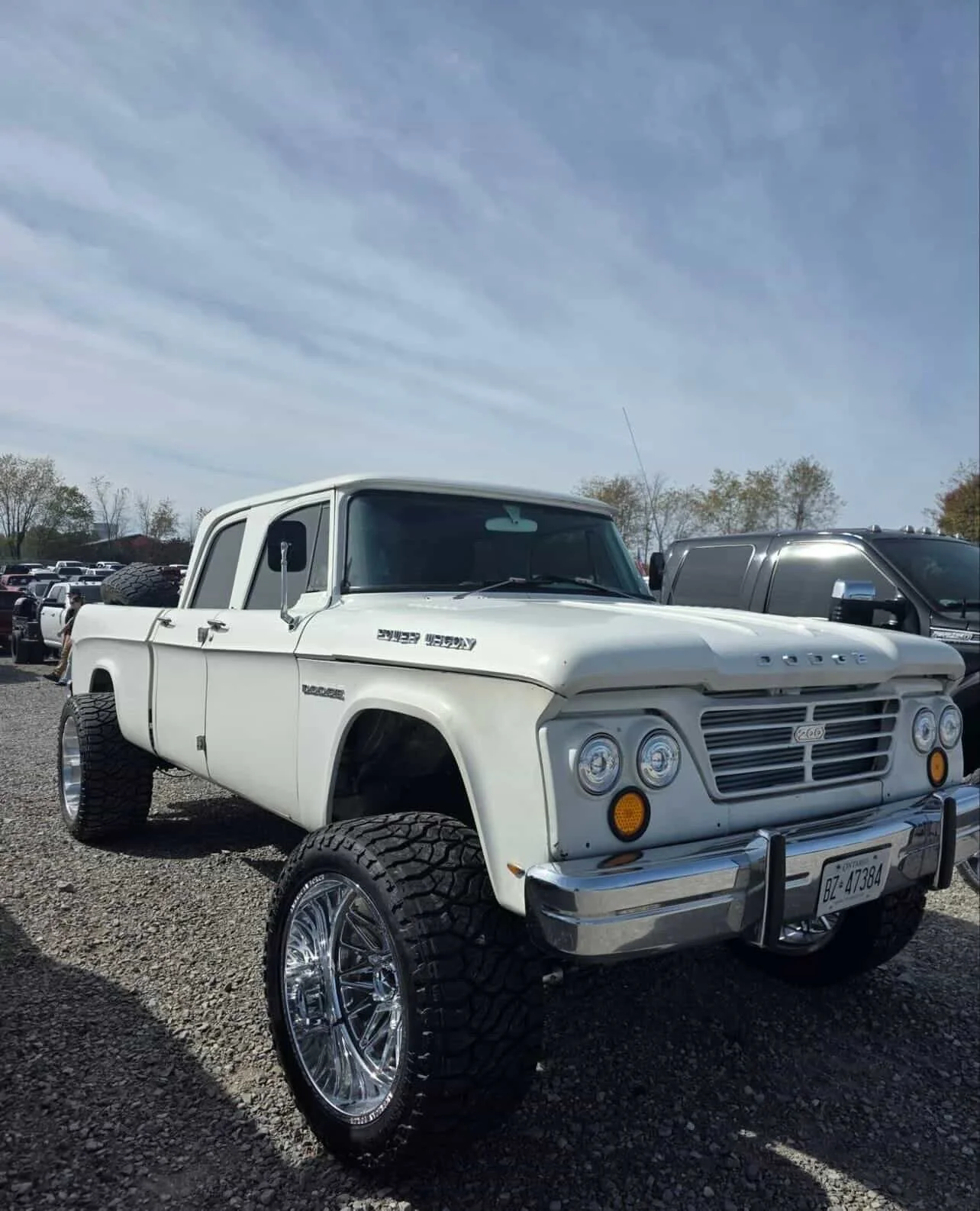 A vintage white Dodge truck with large, shiny aftermarket wheels and off-road tires parked in a car lot with other vehicles under a partly cloudy sky.