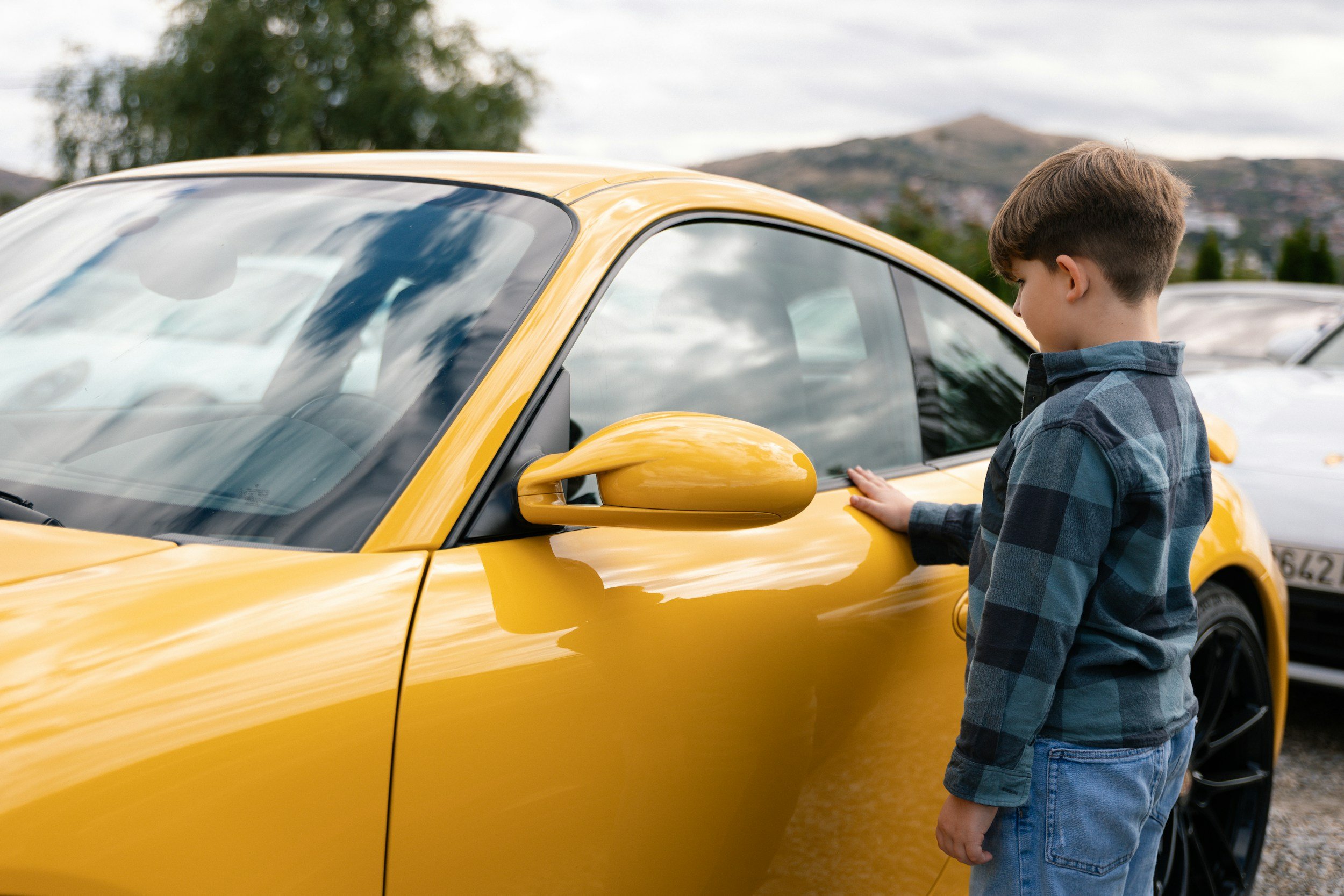 A young boy wearing a blue plaid shirt and jeans is touching the yellow exterior of a sports car, parked outdoors with a background of other cars, trees, and distant hills.