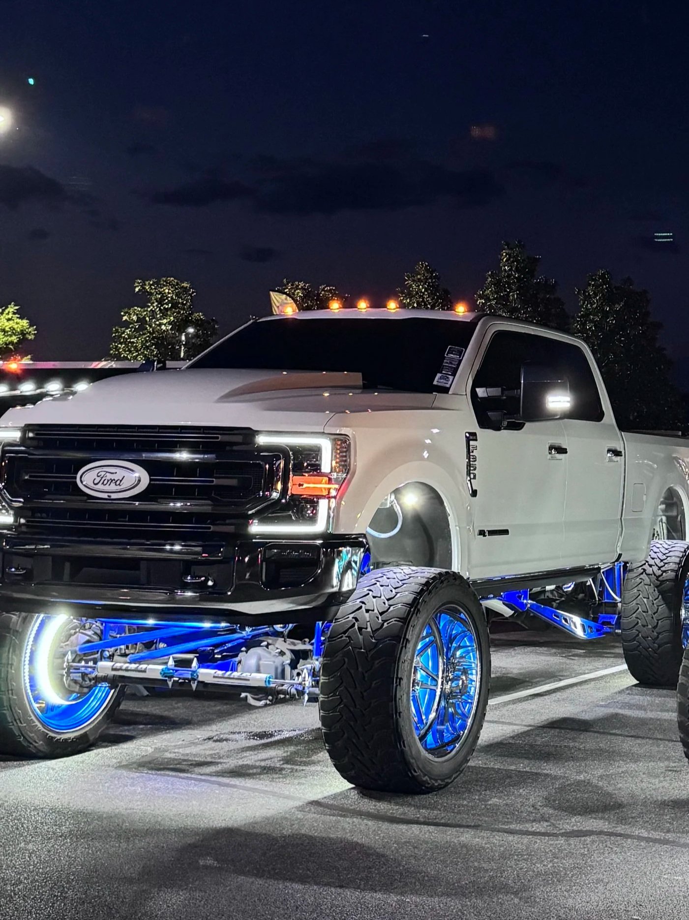 A white lifted Ford truck with large tires and glowing blue rims, decorated with lights, parked in a parking lot at night under a dark sky with trees and a bright moon.