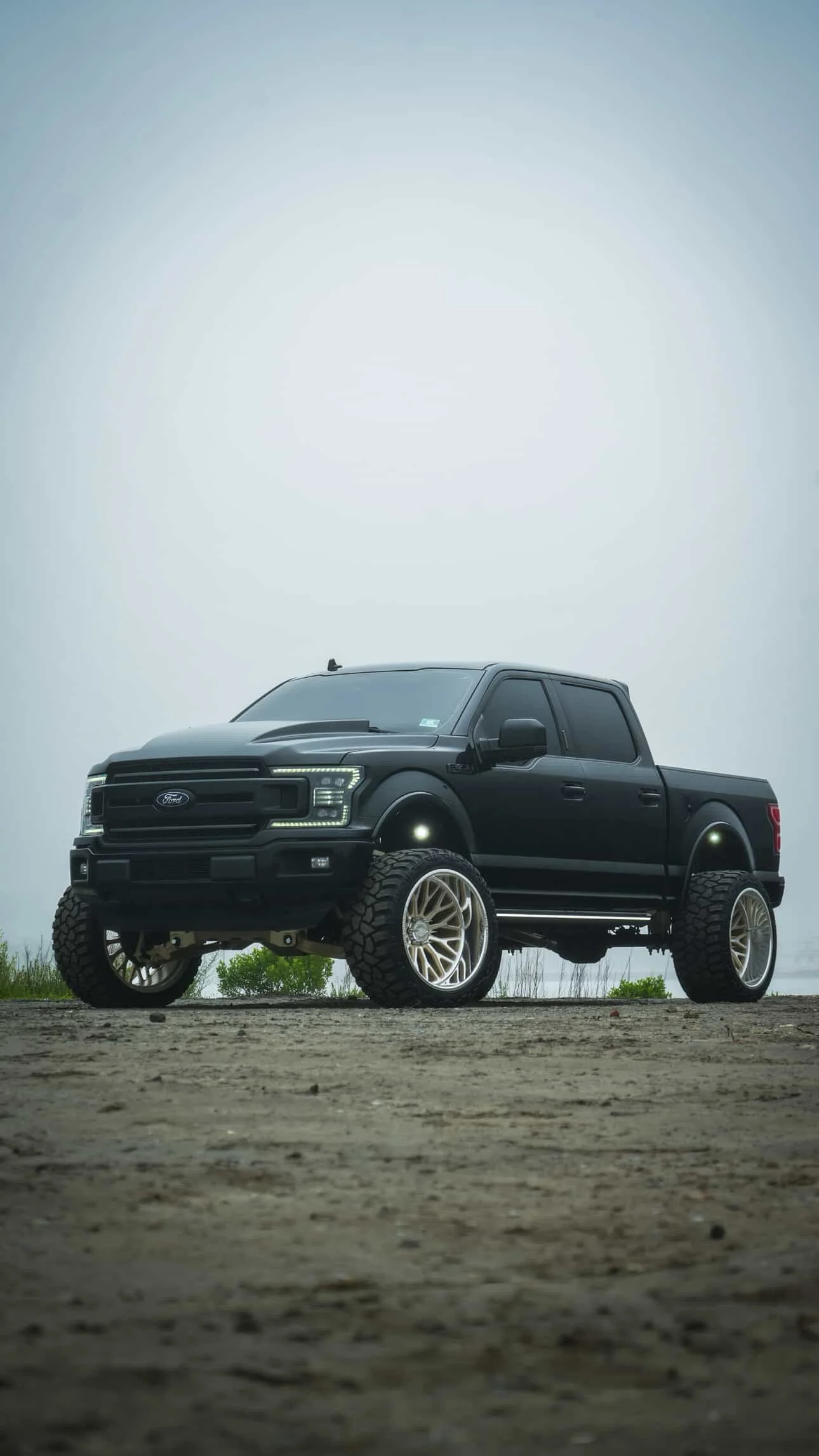 A black Ford pickup truck with large off-road tires and custom rims is parked on a dirt surface under an overcast sky.