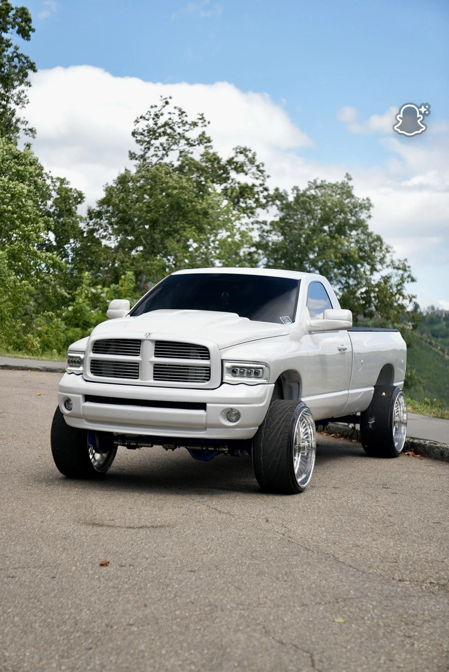 A white pickup truck with large tires parked on a paved road, surrounded by green trees and a partly cloudy blue sky.