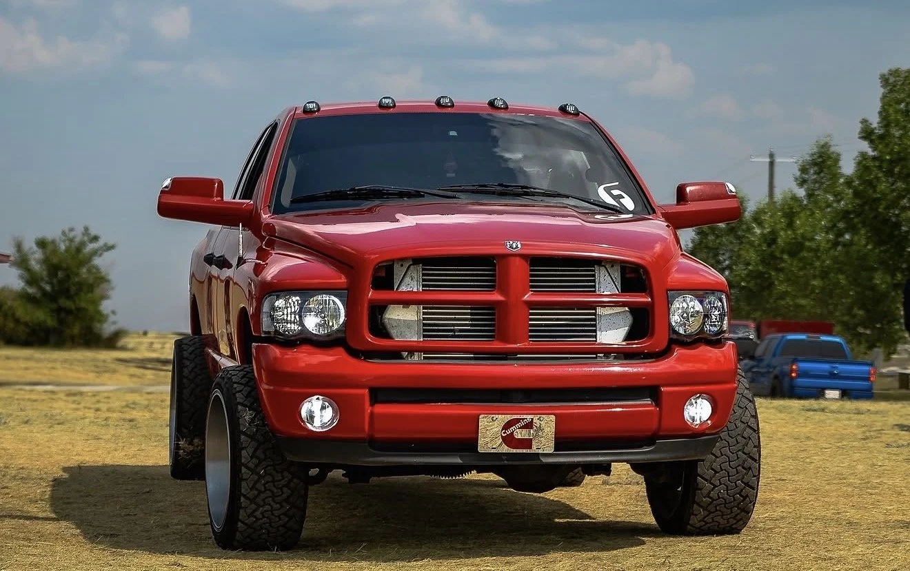 Red pickup truck with a custom grille and front bumper, black off-road tires, parking on a grassy field with other vehicles and trees in the background.