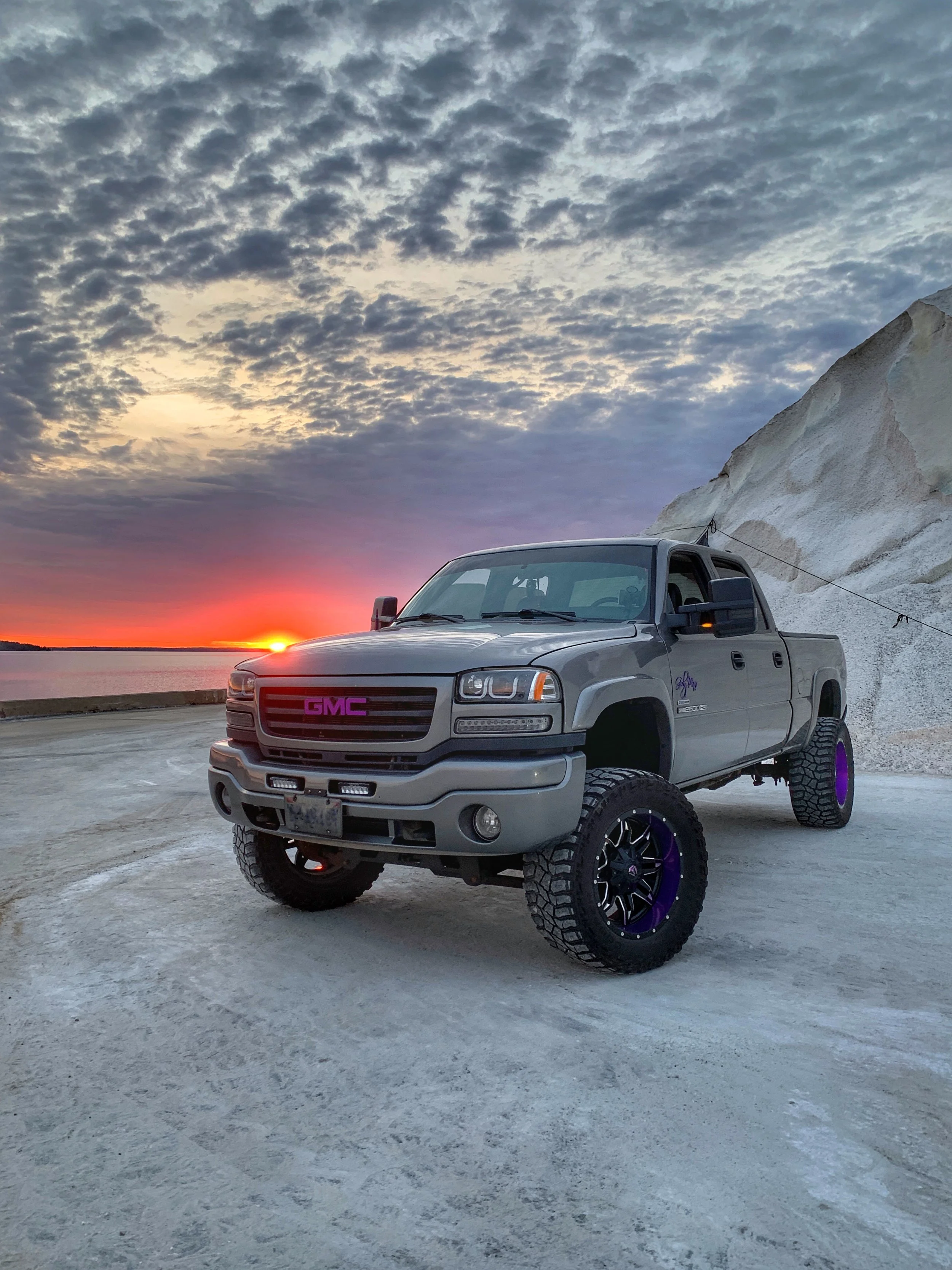 A silver GMC pickup truck with enhanced off-road tires and custom purple accents parked on a flat, light-colored surface near snow-covered terrain, with a view of a sunset sky with scattered clouds.
