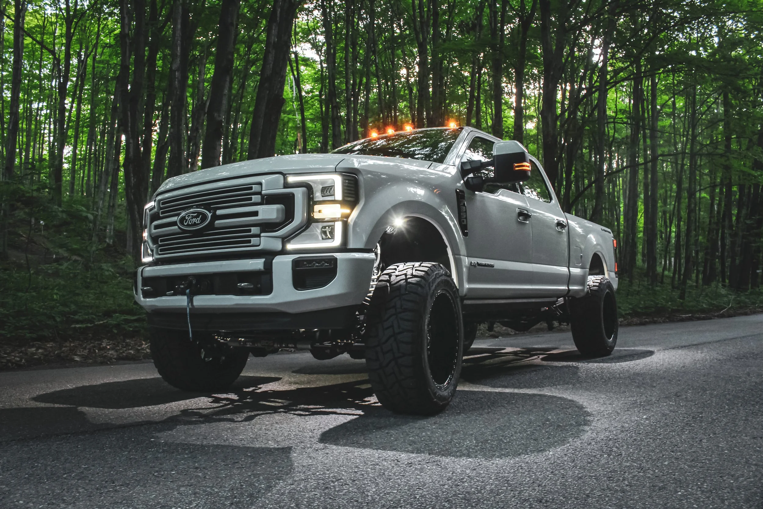 A white Ford pickup truck with deck lights and large off-road tires parked on a road in a dense green forest.