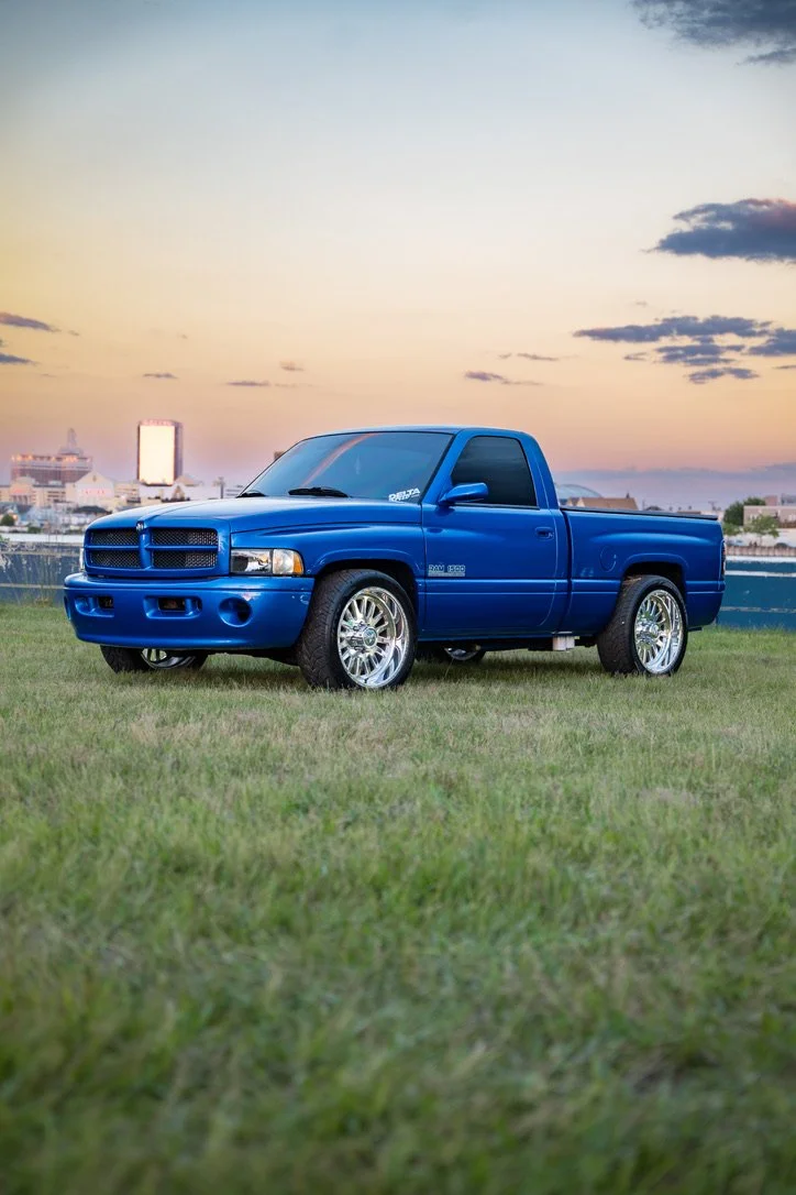 A blue pickup truck parked on grass with a city skyline and sunset in the background.