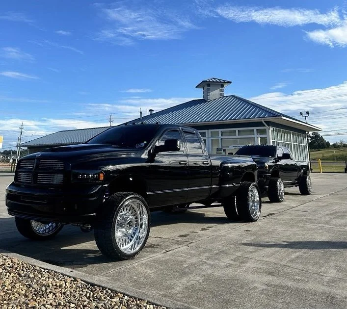 Black pickup trucks with large shiny wheels parked outside a building with a blue metal roof and a small tower on top during daytime.