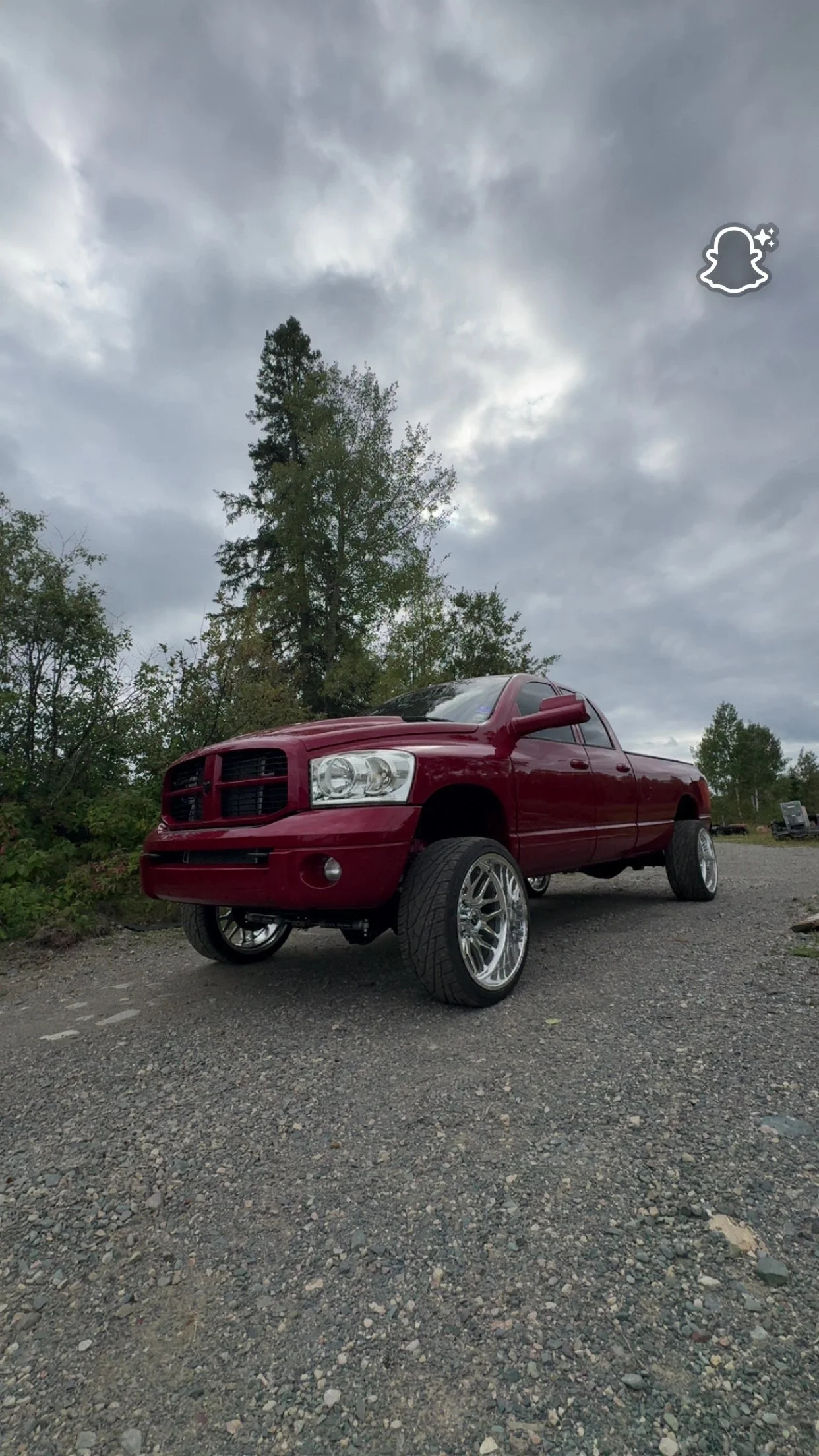 Red pickup truck with custom rims parked on a gravel lot under a cloudy sky, surrounded by trees.