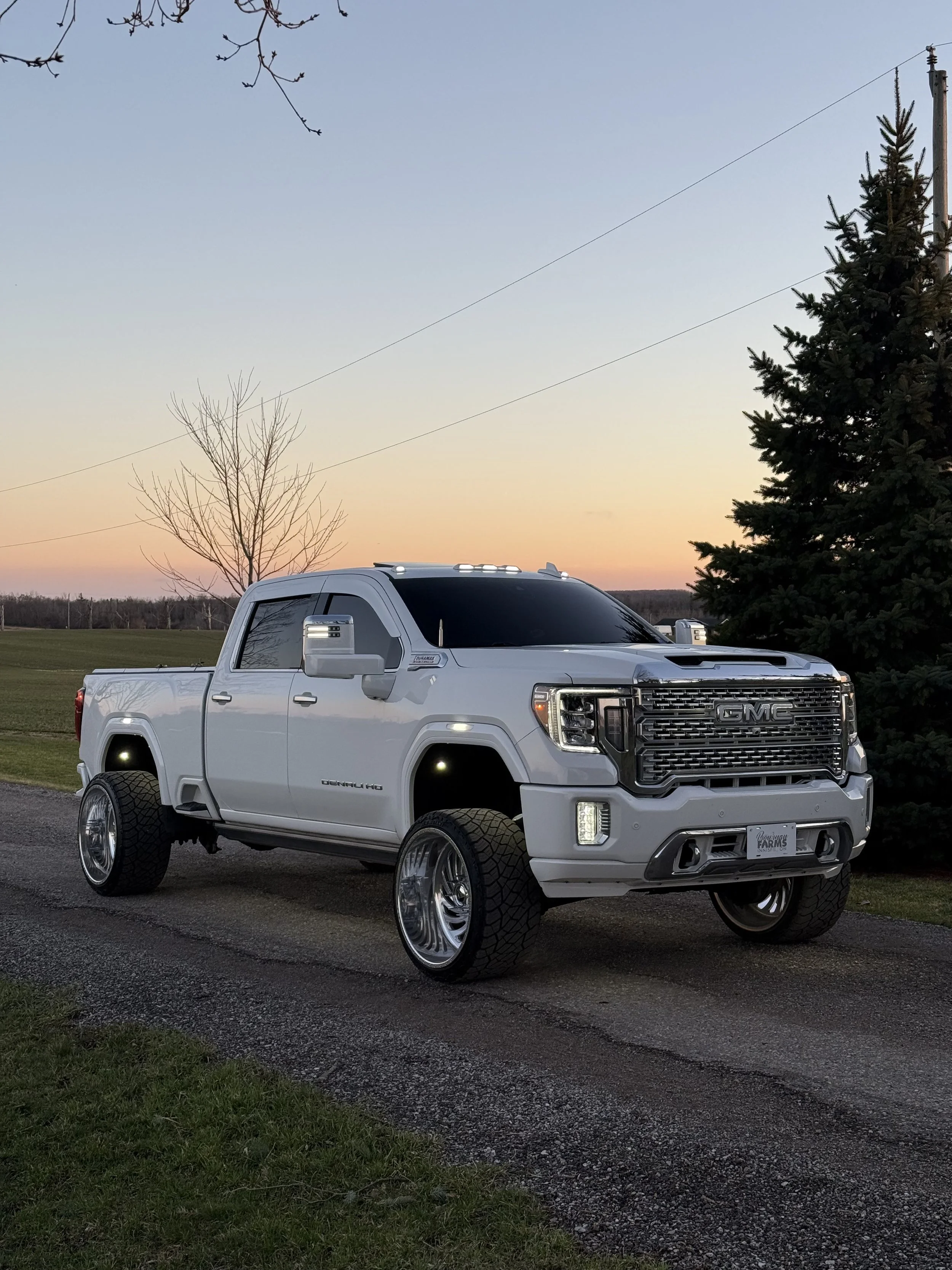 A white GMC pickup truck with oversized wheels parked on a gravel road at sunset, with leafless trees and a large evergreen tree in the background.