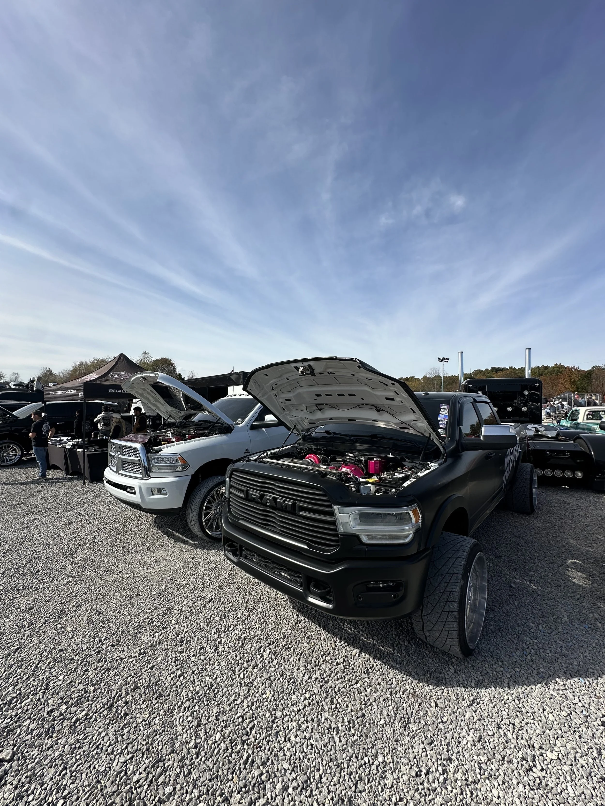 Cars on display at an outdoor car show, with their hoods open showcasing engines, and a crowd of people around. Clear sky and tents visible in the background.