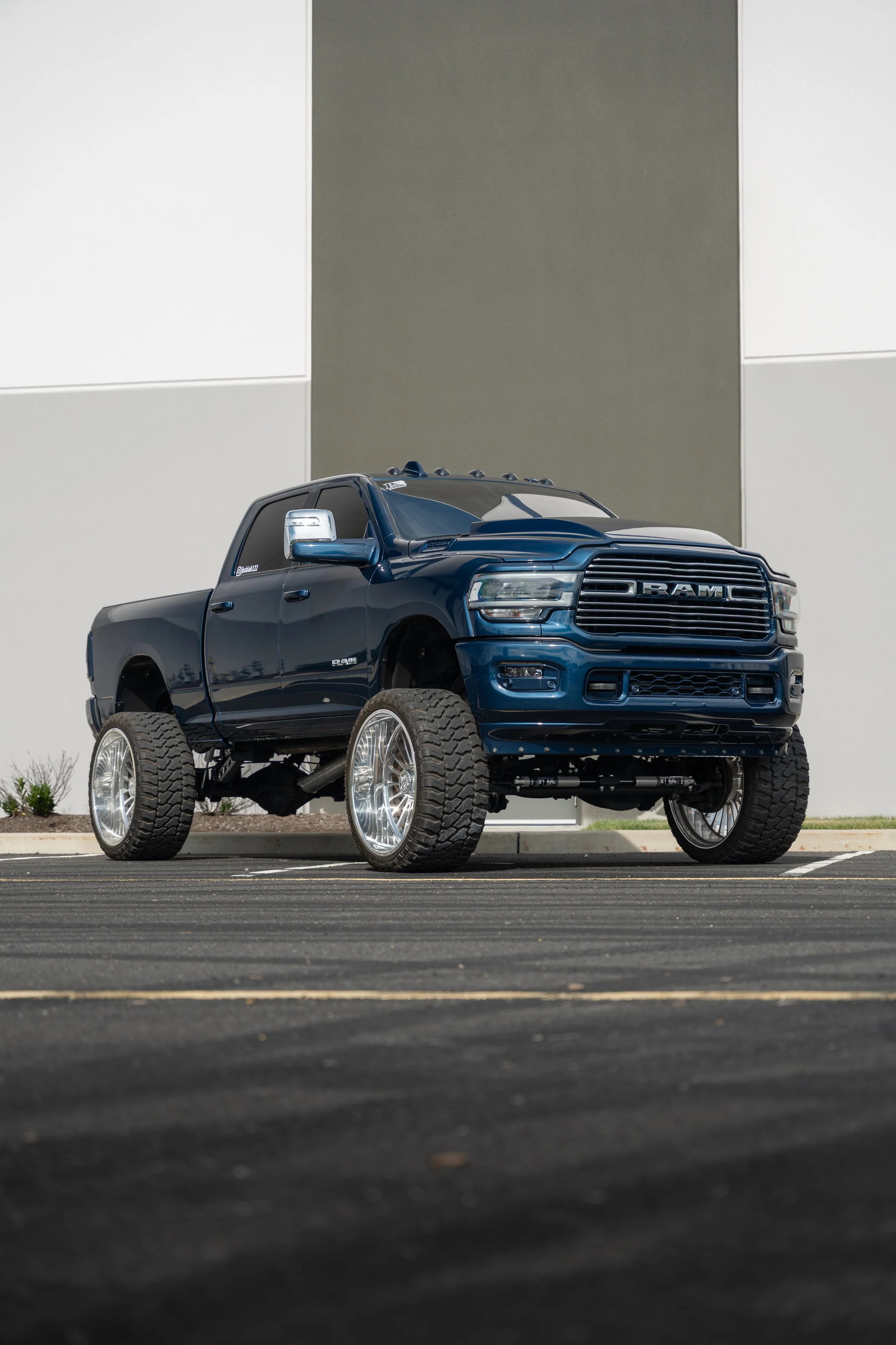 A model of a blue RAM pickup truck with large tires parked in a parking lot in front of a building with white and grey walls.