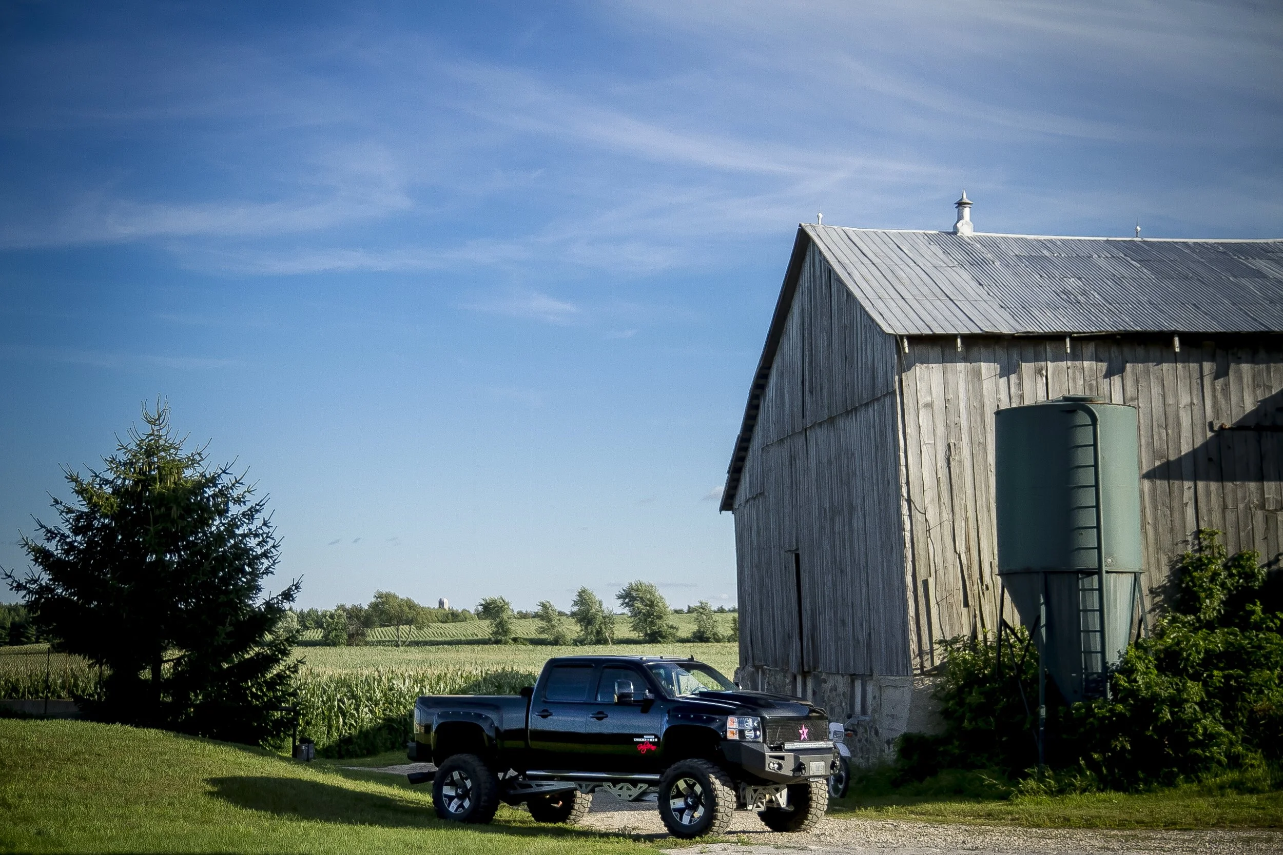 A black pickup truck parked on a dirt driveway near a wooden barn with corrugated metal roof on a sunny day. Green fields and trees are visible in the background under a blue sky with some clouds.