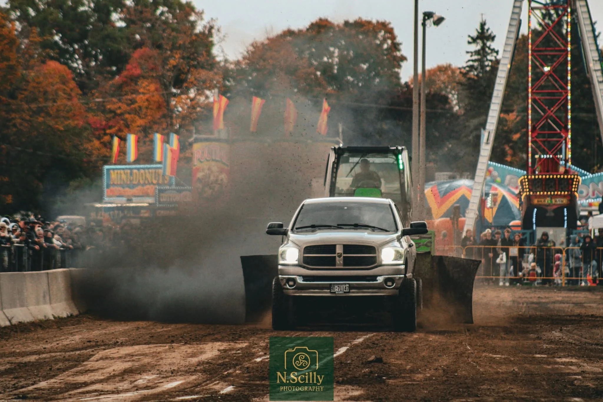 A race car speeds down a dirt track at a fairground, creating dust in the air. In the background, a crowd watches the race and amusement rides, including a large Ferris wheel and a roller coaster, with colorful flags and signs visible. The setting su