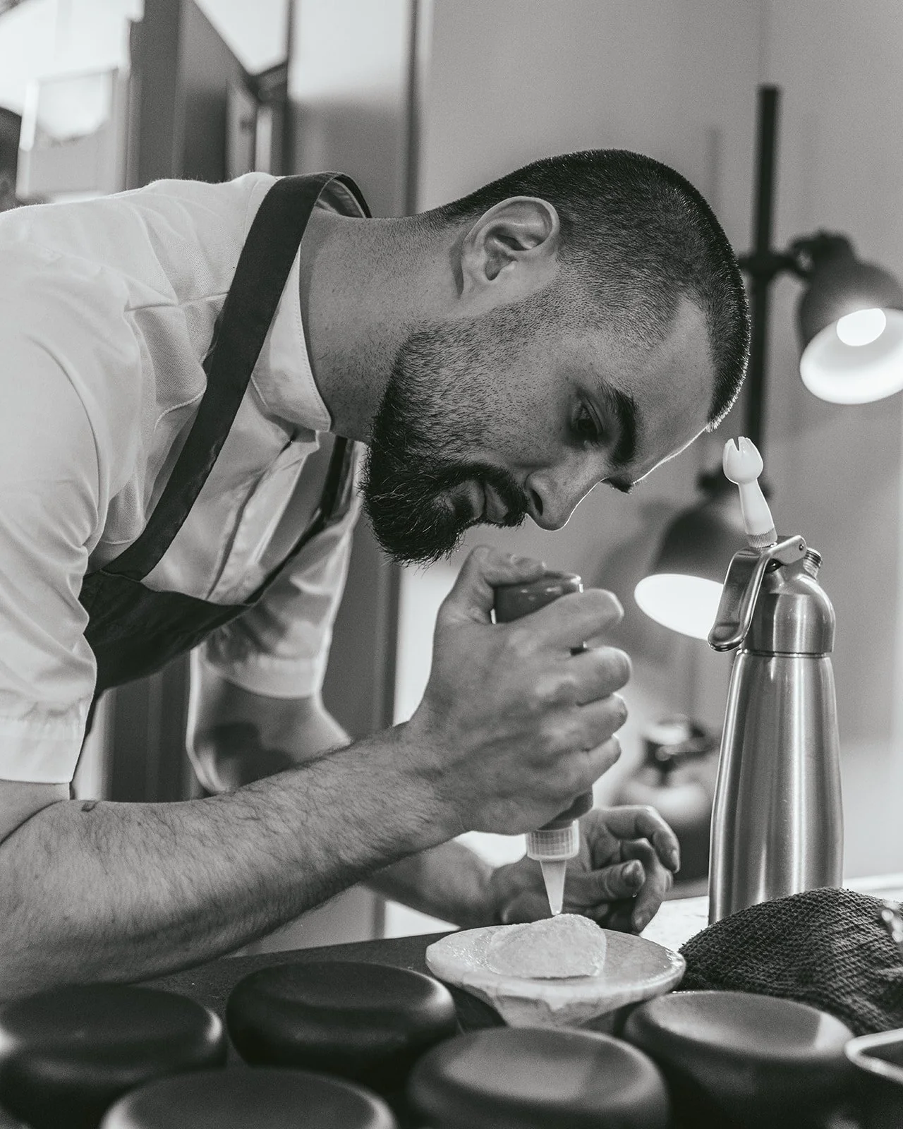 A man with a beard and short hair, wearing a white shirt and black apron, is using a dropper to place a liquid onto a stone or mineral piece on a desk in a well-lit room with framed pictures and a lamp in the background.
