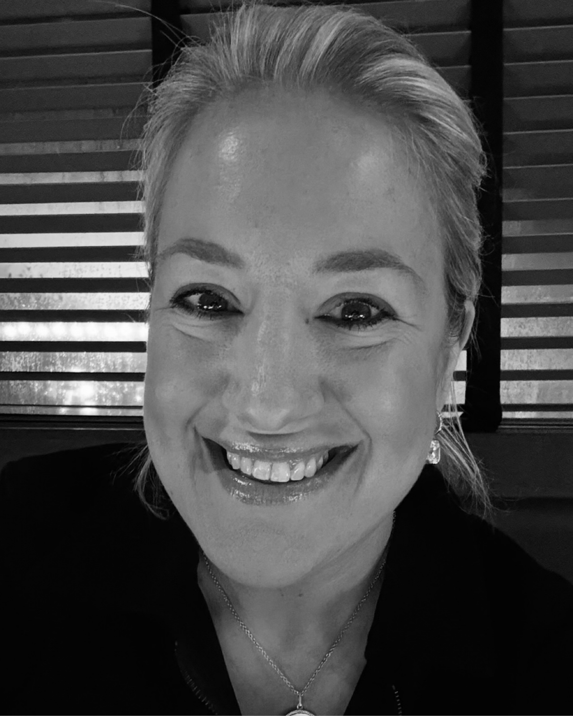 Close-up black-and-white portrait of a smiling woman with short, light-colored hair, wearing earrings and a necklace, with a background of horizontal wooden slats.