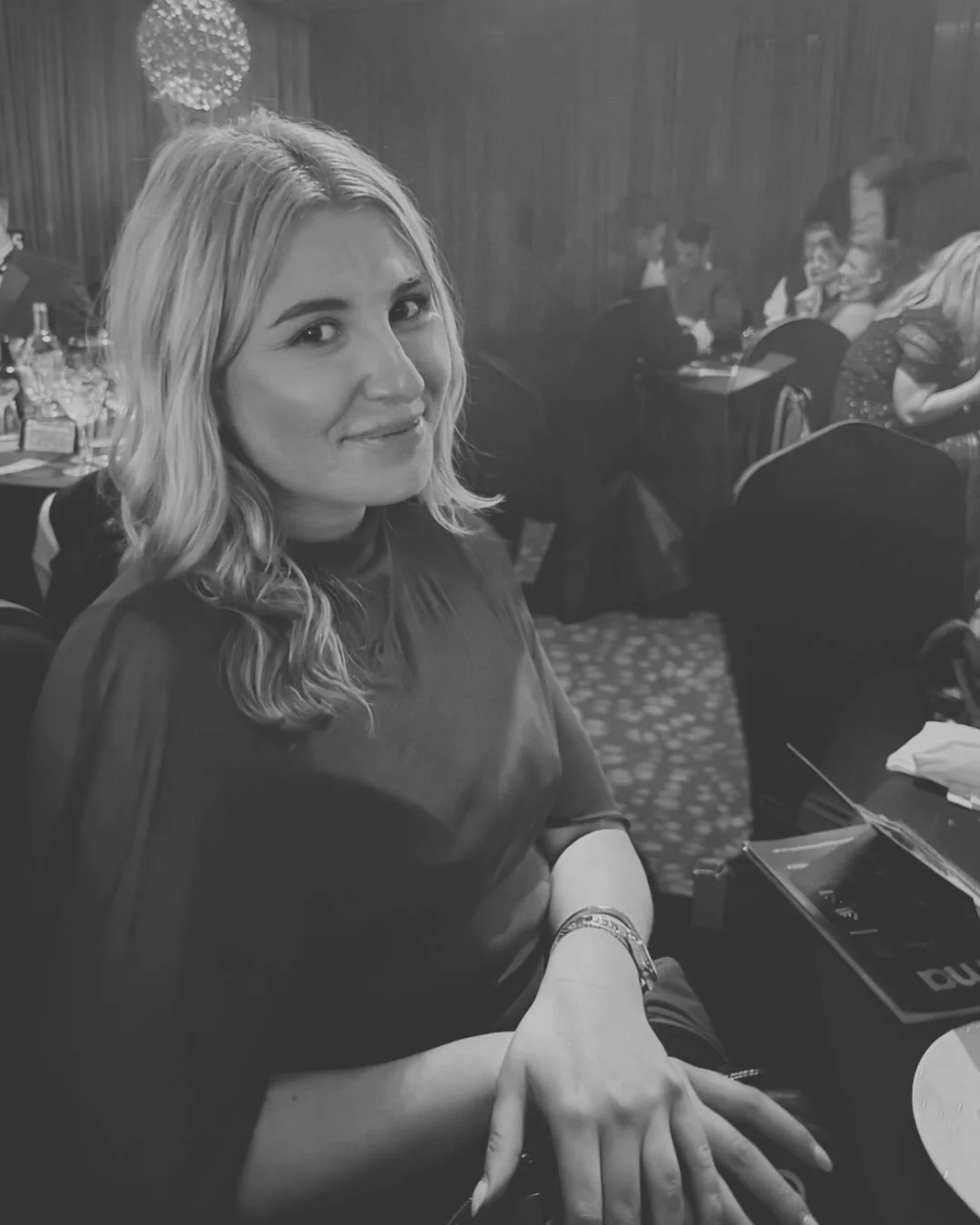 A woman with blonde, wavy hair smiling at the camera in a restaurant or cafe setting, with other people seated in the background.
