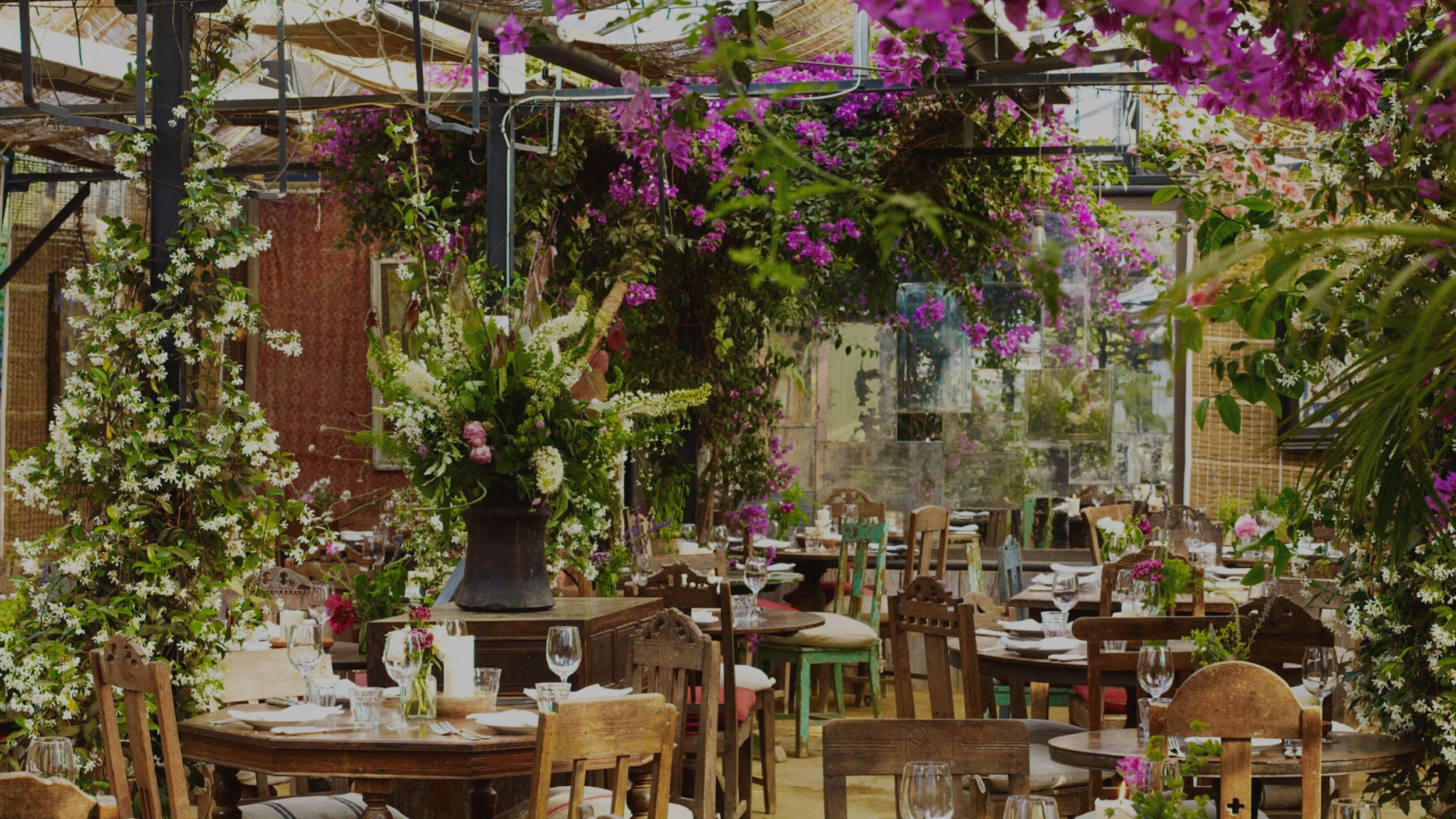 A cozy restaurant interior bursting with colorful flowering plants and greenery, featuring rustic wooden tables and chairs set for dining.