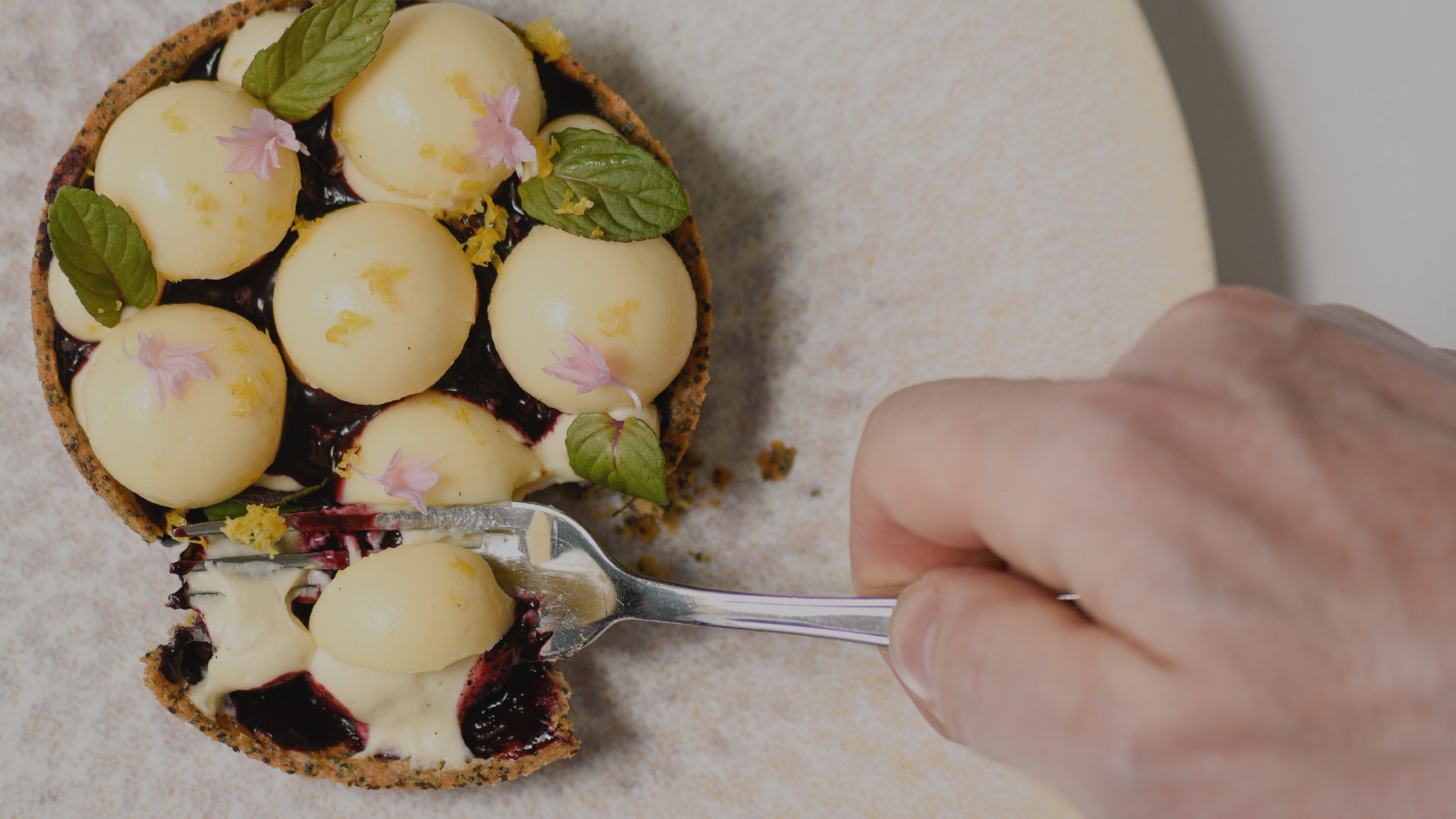 Top view of a person cutting into a fruit tart with yellow cream and purple and green garnishes on a beige surface.