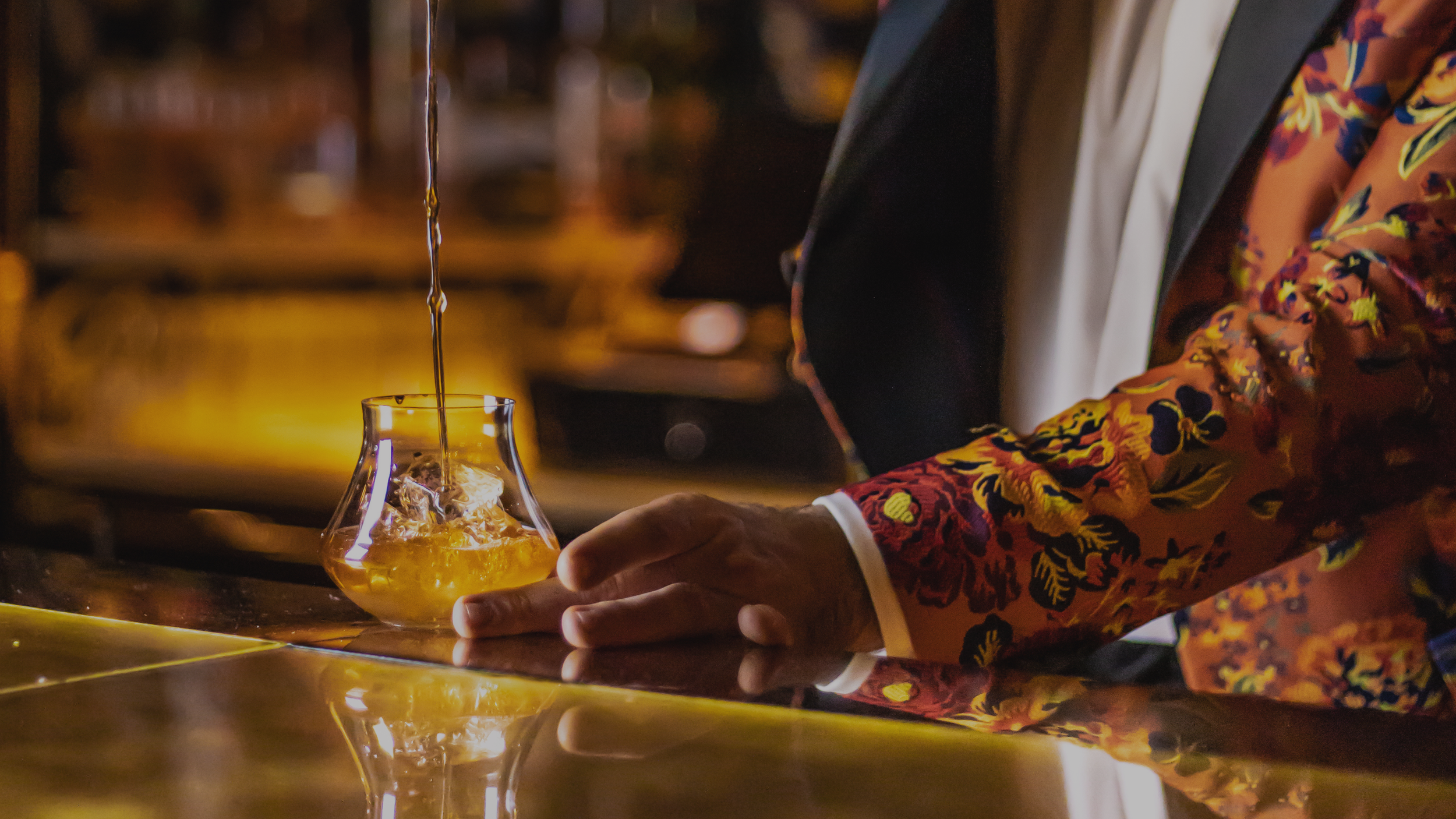 A man pouring liquor into a glass with ice on a bar counter at night, wearing a colorful floral blazer.