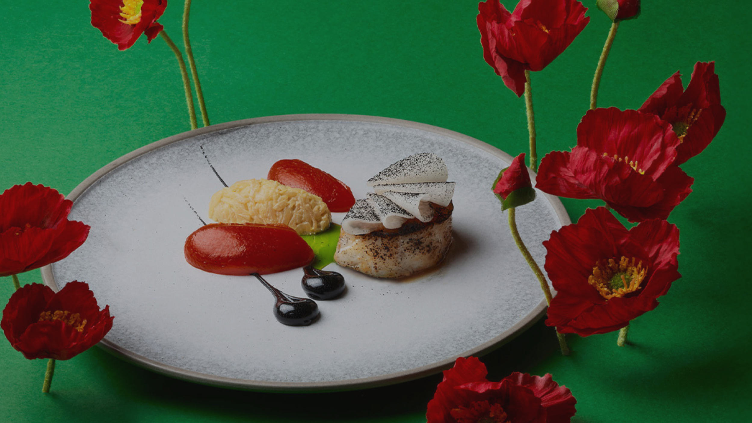 A plated dessert with a small cake topped with cream, two dollops of red sauce, a scoop of pale yellow ice cream, and three small droplets of dark sauce, garnished with a green leaf, surrounded by red poppies on a green background.