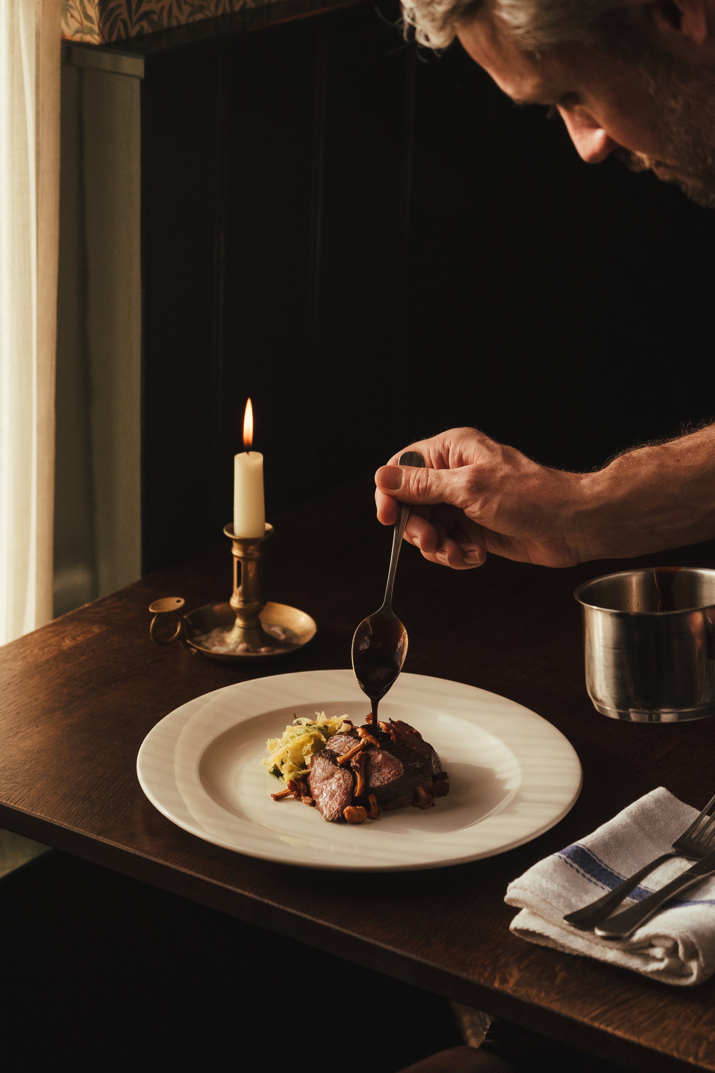 Man pouring dark gravy over a plated dish of steak, mashed potatoes, and vegetables at a restaurant table with a lit candle as ambient lighting.