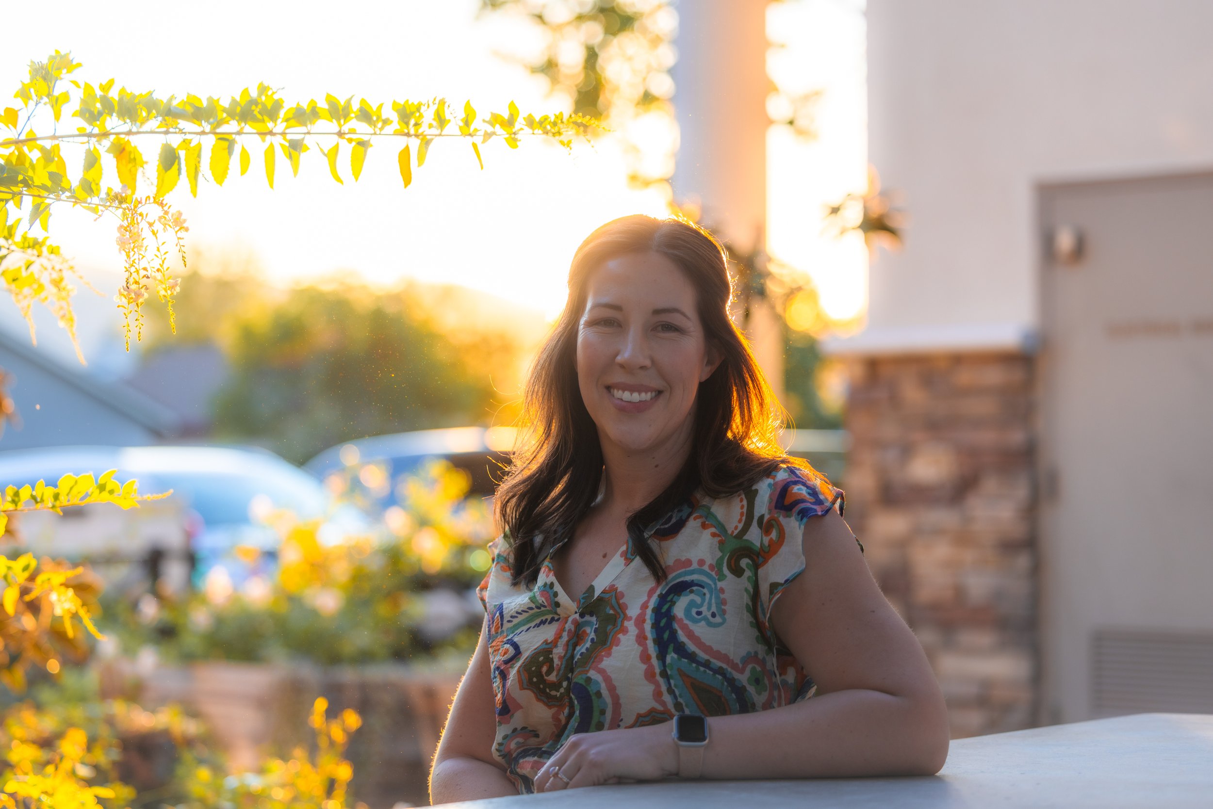 A woman with long dark hair smiling outdoors during sunset. She is wearing a colorful, patterned dress and has a black watch on her left wrist. There are yellow green leaves above her and a blurred background with cars and buildings.