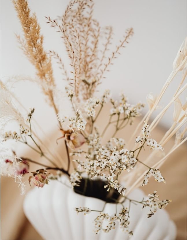 A white vase holding dried flowers and grasses, including pampas grass and small white blossoms, on a light-colored surface.