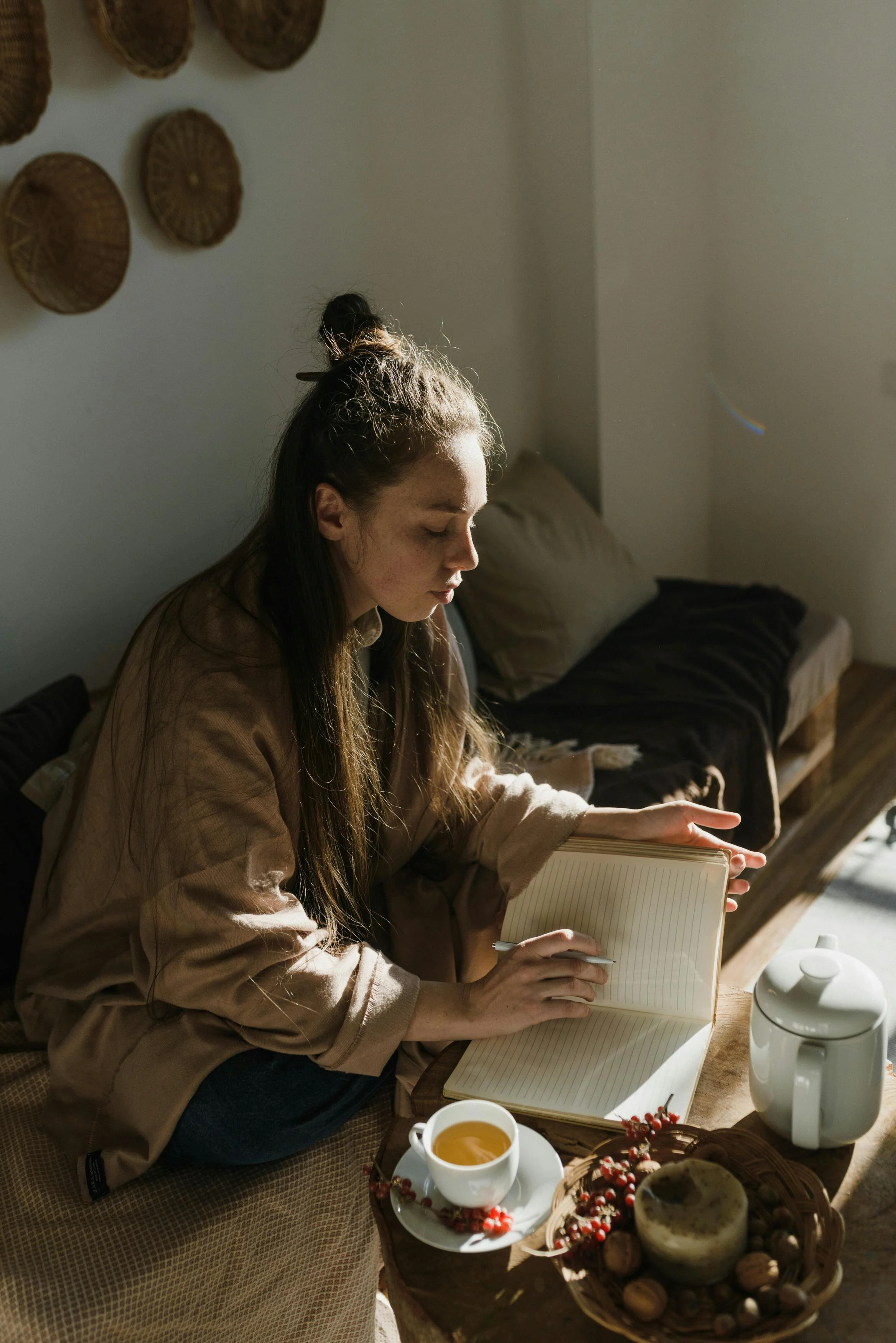A young woman with long hair tied up in a bun, sitting on a bed, reading a notebook. She is wearing a brown long-sleeve shirt. In front of her on a wooden table are a white teapot, a cup of tea, a bowl with berries, some muffins, and nuts. The room has a cozy, warm ambiance with soft lighting and wicker wall decor.