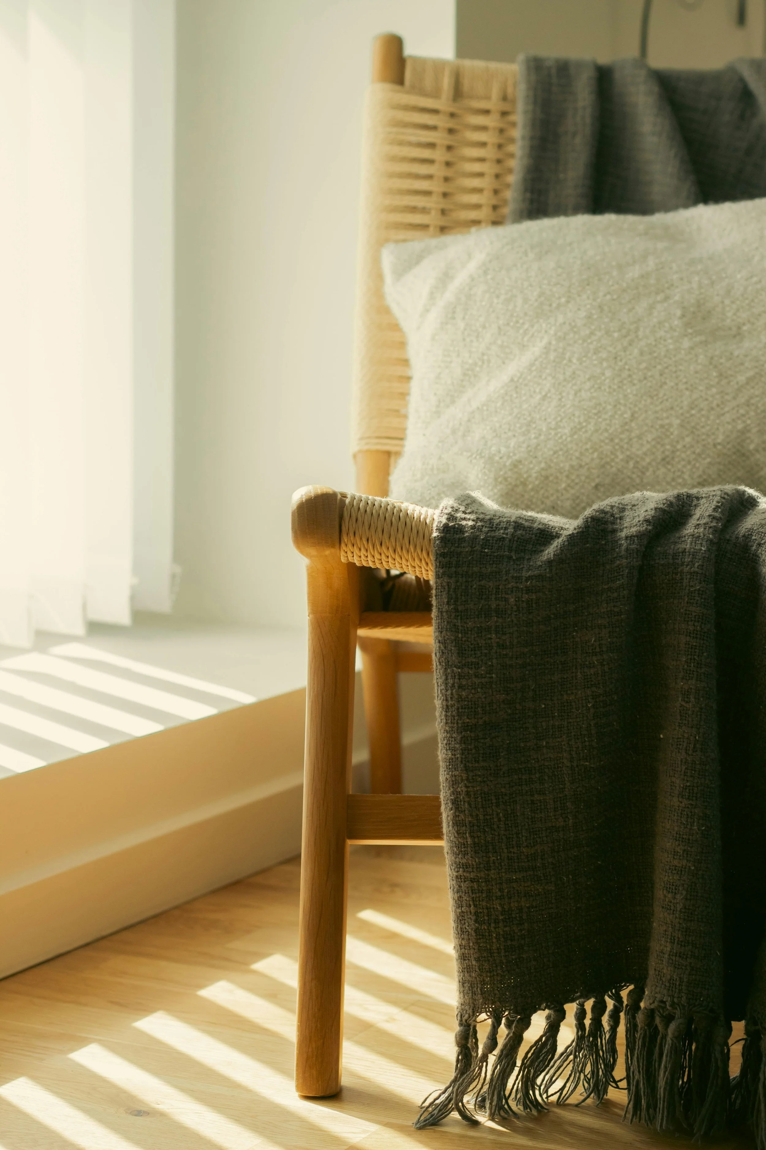 Sunlight streams through a window, casting shadows on the floor next to a wooden chair with woven rattan backrest. A dark gray scarf with tassels hangs over the chair's arm, and a beige cushion rests on the seat.
