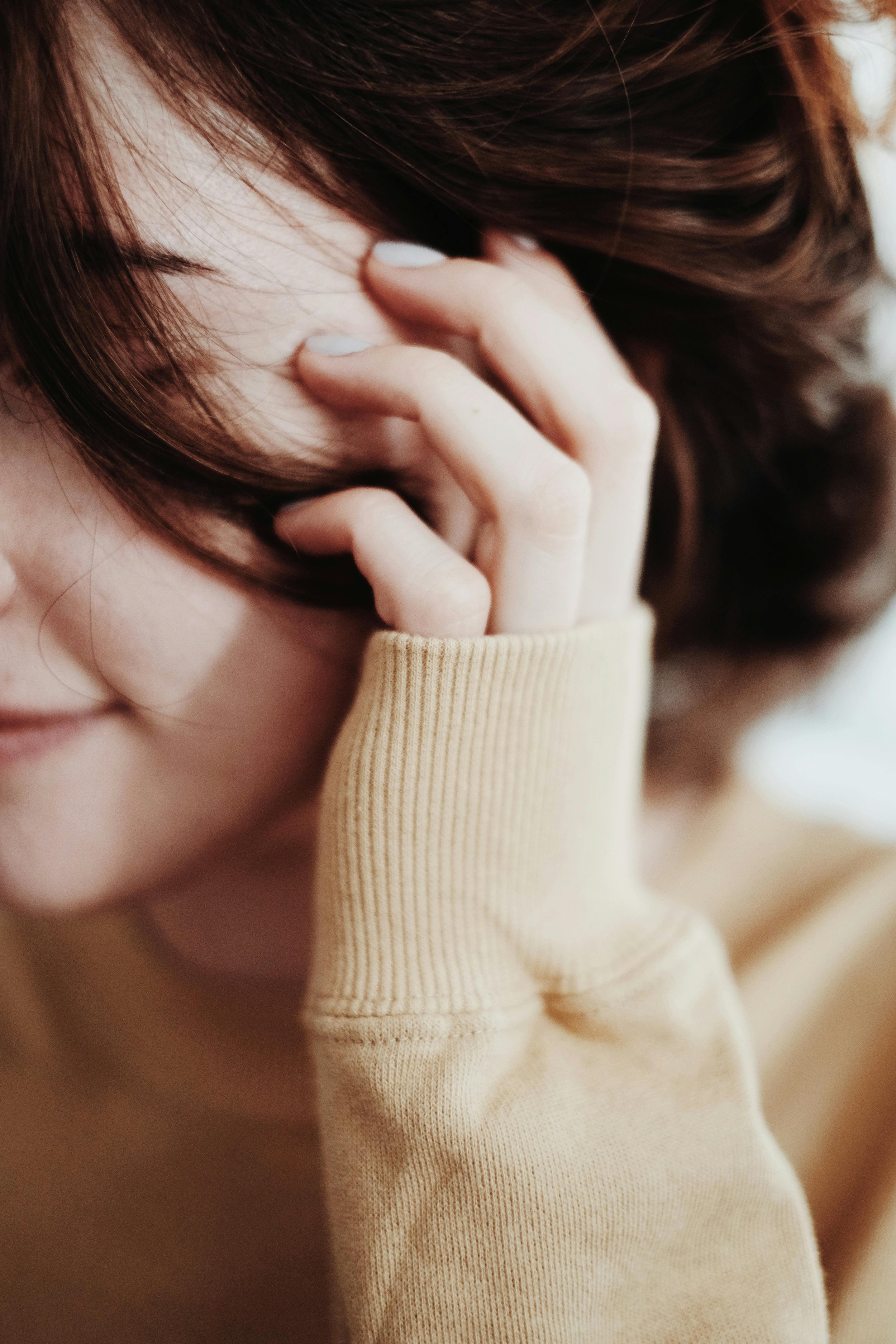 Close-up of a person with long brown hair, partially covering their face, touching their forehead with their hand, wearing a beige long-sleeve shirt.