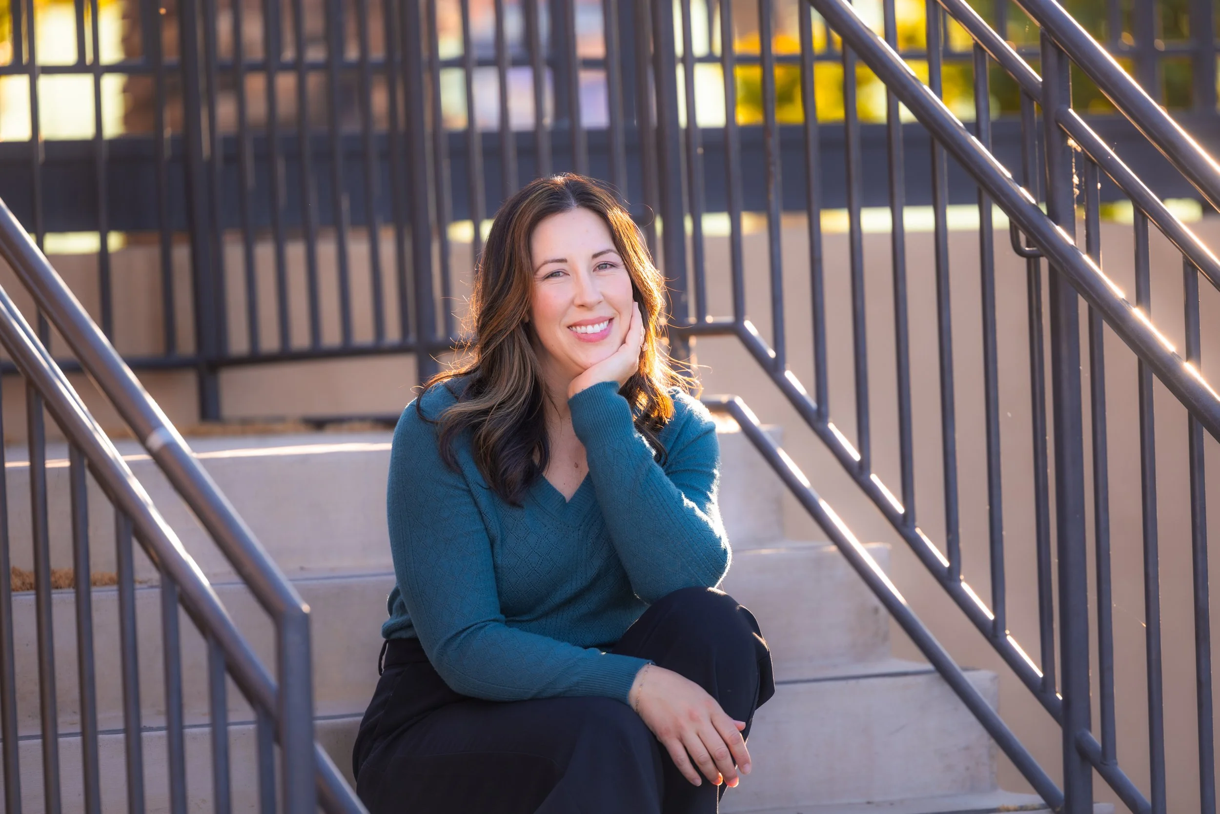 A woman with long dark hair sitting on concrete stairs next to a metal railing, smiling and resting her chin on her hand in warm sunlight.