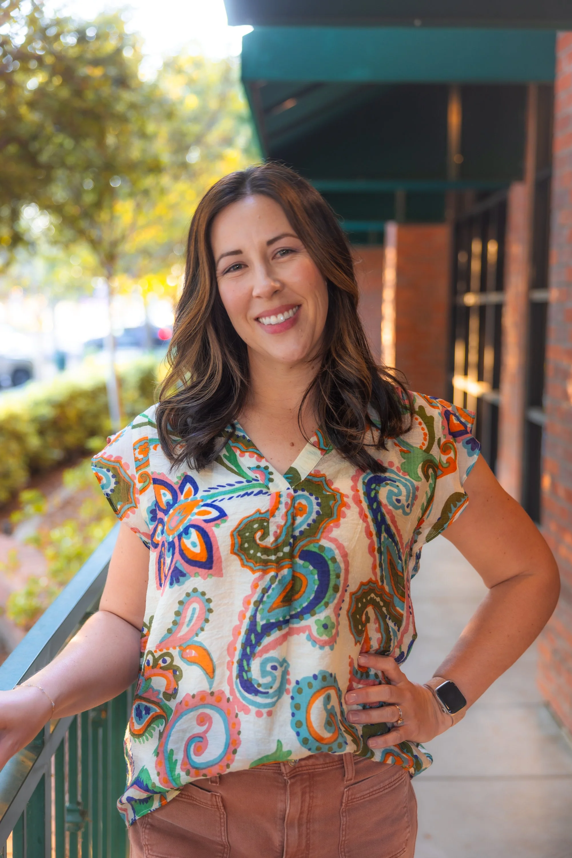 Woman with shoulder-length brown hair smiling outdoors, wearing a colorful paisley patterned blouse, brown pants, standing next to a green railing against a background of trees and brick building.