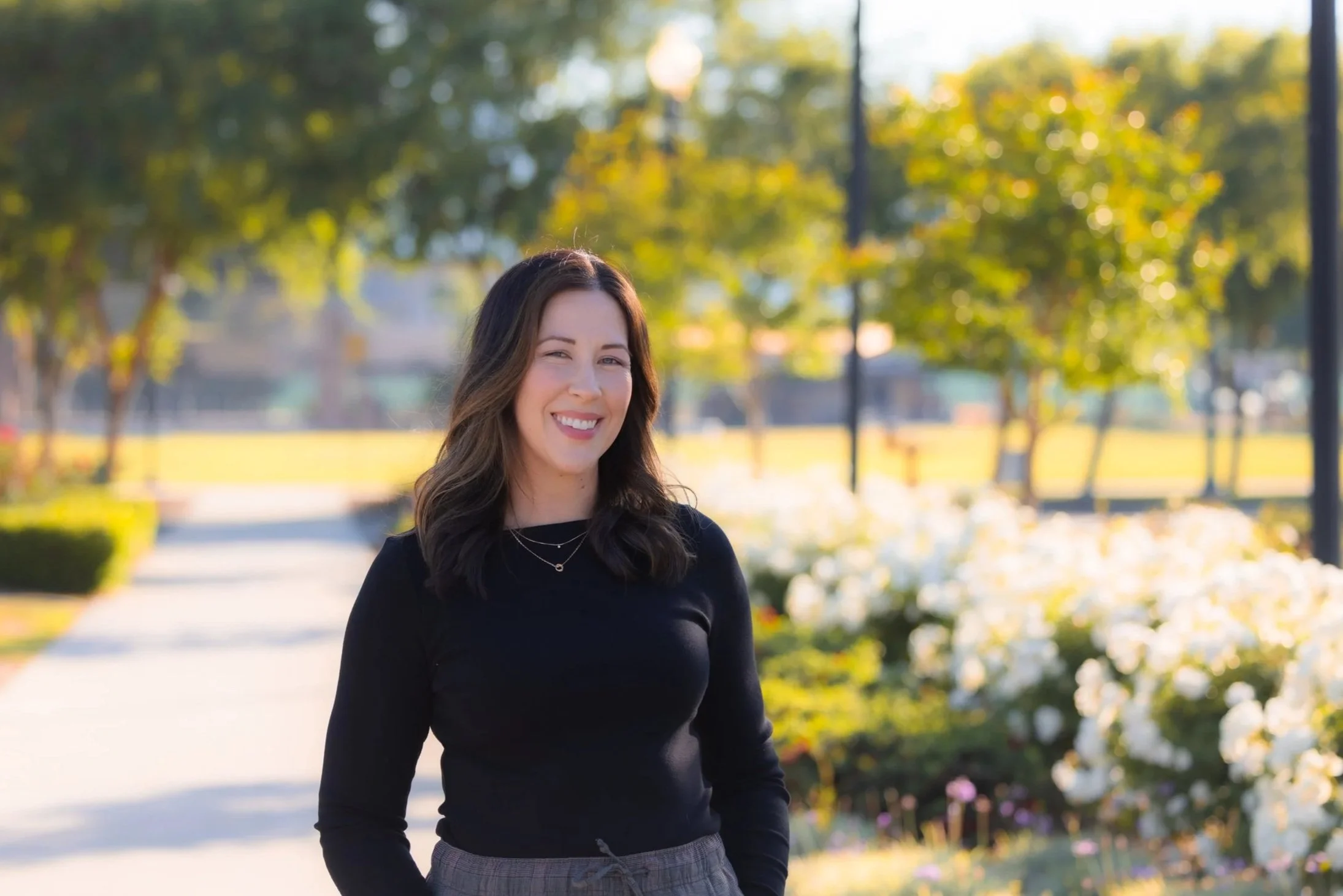 A woman with dark brown hair smiling outdoors in a park during fall, wearing a black top and gray checkered pants, with trees and greenery in the background.