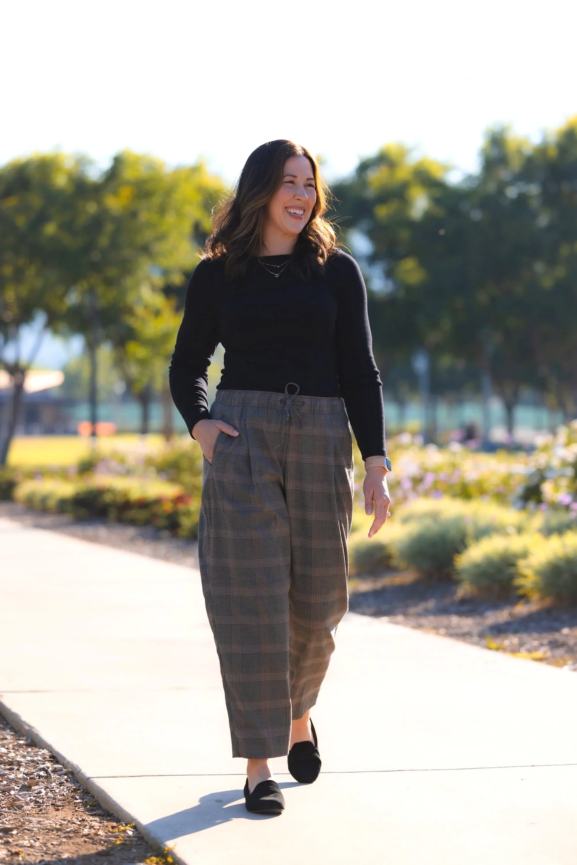 A woman with shoulder-length brown hair, wearing a black long-sleeve top and plaid wide-leg pants, is walking outdoors on a sunny day, smiling and looking to her right with trees and a park in the background.