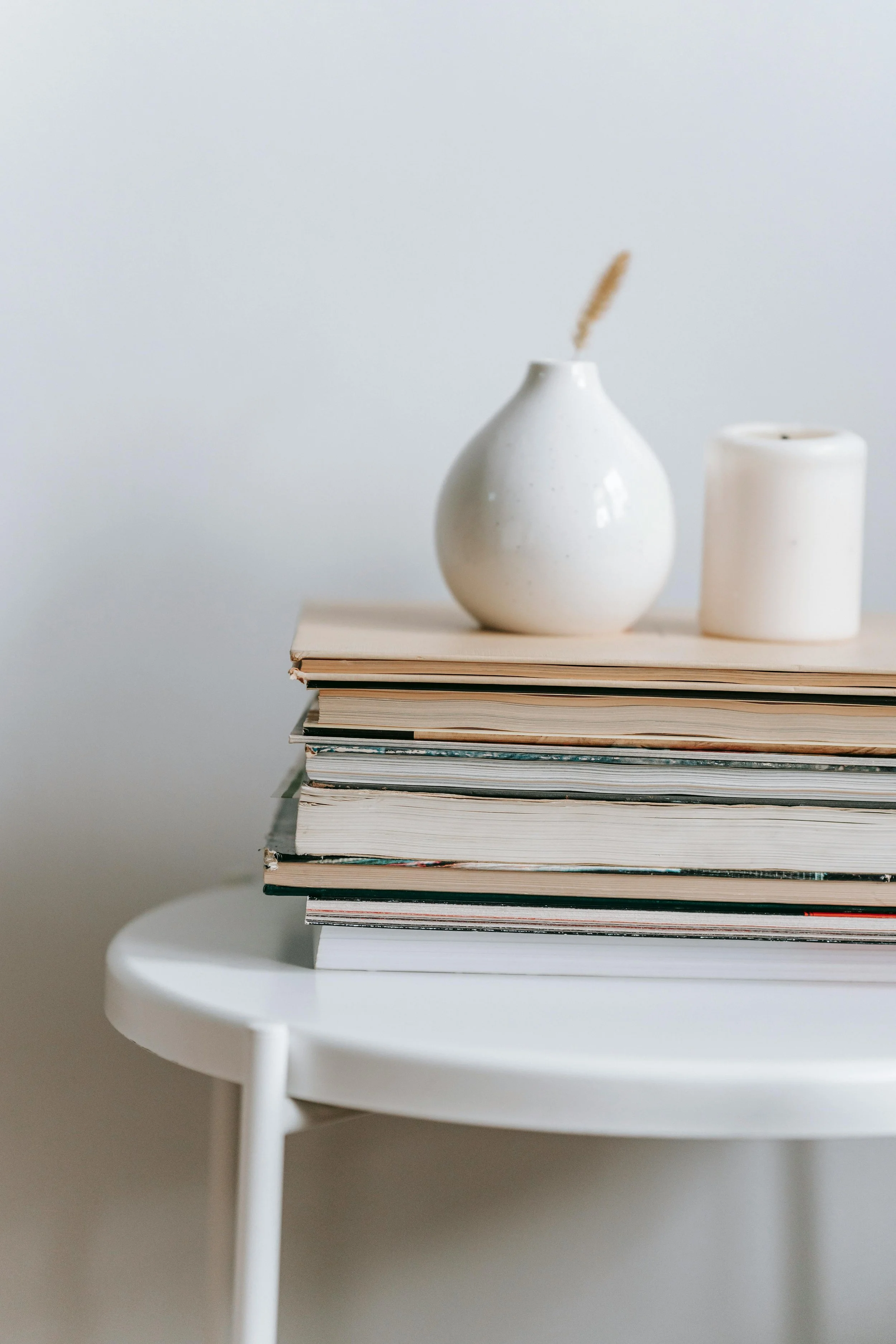 Stack of books on a white round table with three white vases on top, the largest vase has a dried wheat stalk inside, the background is light gray.