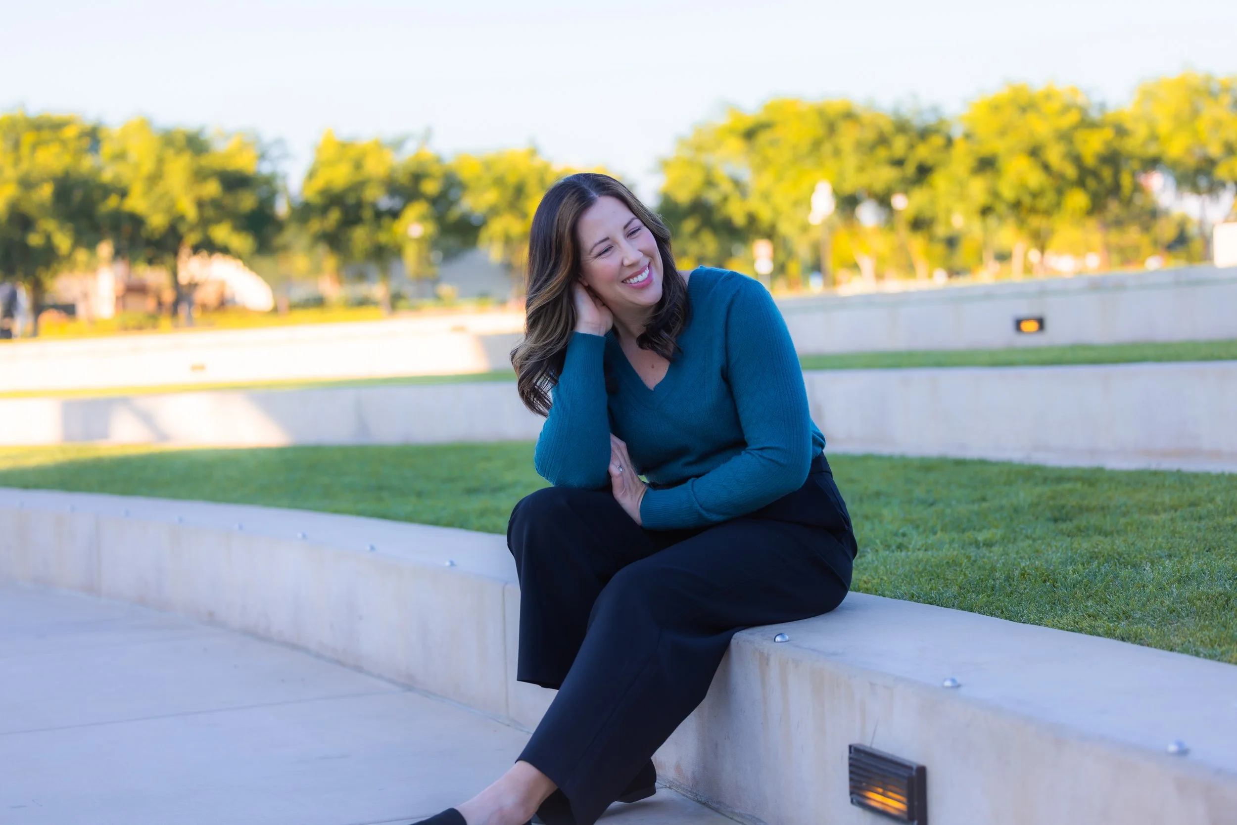 A woman with brown hair, wearing a teal long-sleeve shirt and black pants, sits on a concrete ledge outdoors, smiling with her eyes closed, in a park with green grass, trees, and a clear sky.