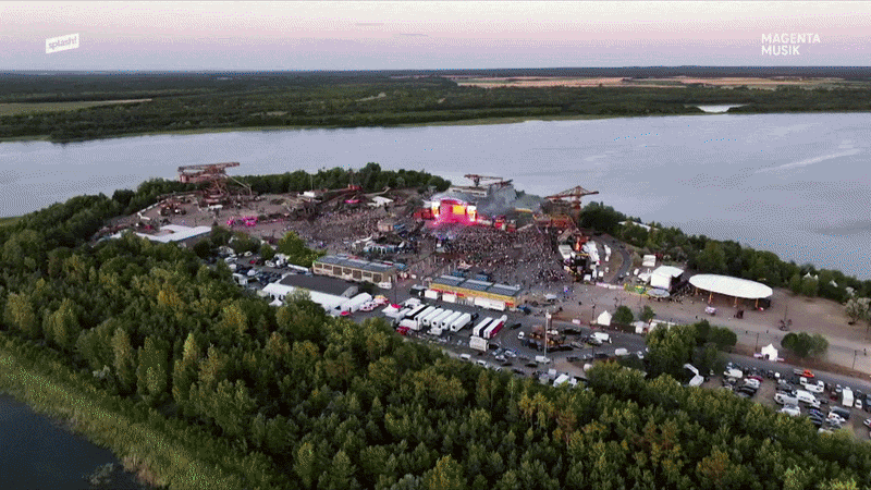Luftaufnahme eines Musikfestivals am Fluss, umgeben von Bäumen, mit Bühnen, Zelten und Menschenmenge, bei Sonnenuntergang.