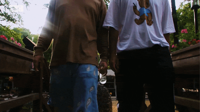Zwei Personen stehen auf einer Holzbrücke im Freien, mit Bäumen und Pflanzen im Hintergrund.