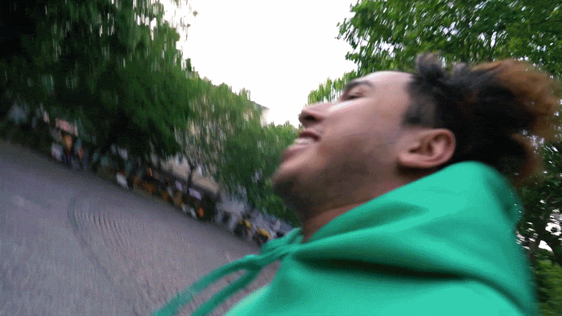 Ein Mann läuft in einem Park oder an einer Straße, umgeben von grünen Bäumen, trägt einen grünen Pullover und hat braune, lockige Haare.