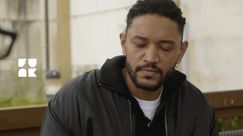 Ein Mann mit kurzen, lockigen Haaren und Einohrschmuck sitzt vor einem Hintergrund mit einem Fenster, sieht nach unten, wirkt nachdenklich.