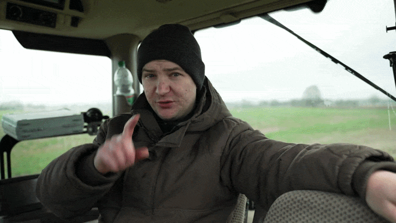 Ein Mann in warmer Kleidung und Mütze sitzt in einem Traktor und macht eine Geste mit der Hand, während er in die Kamera schaut.