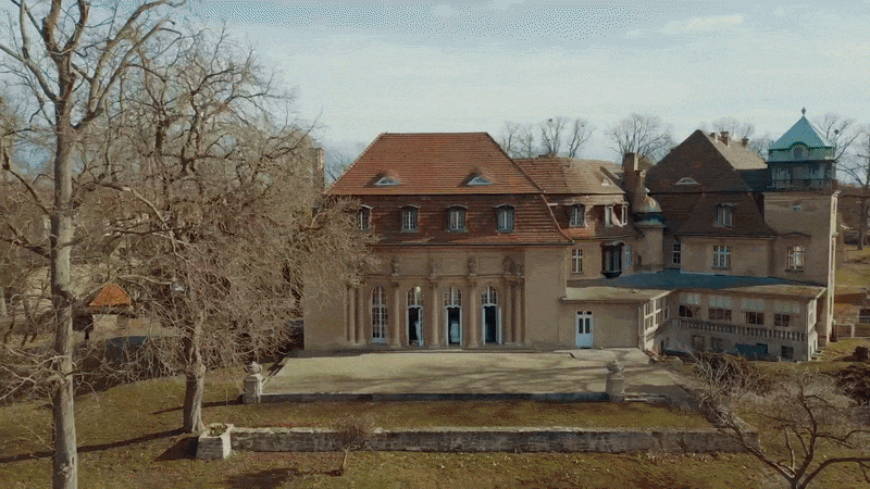 Ein großes historisches Schloss oder Herrenhaus mit rotem Dach, umgeben von Bäumen ohne Blätter, im Frühling oder Winter.
