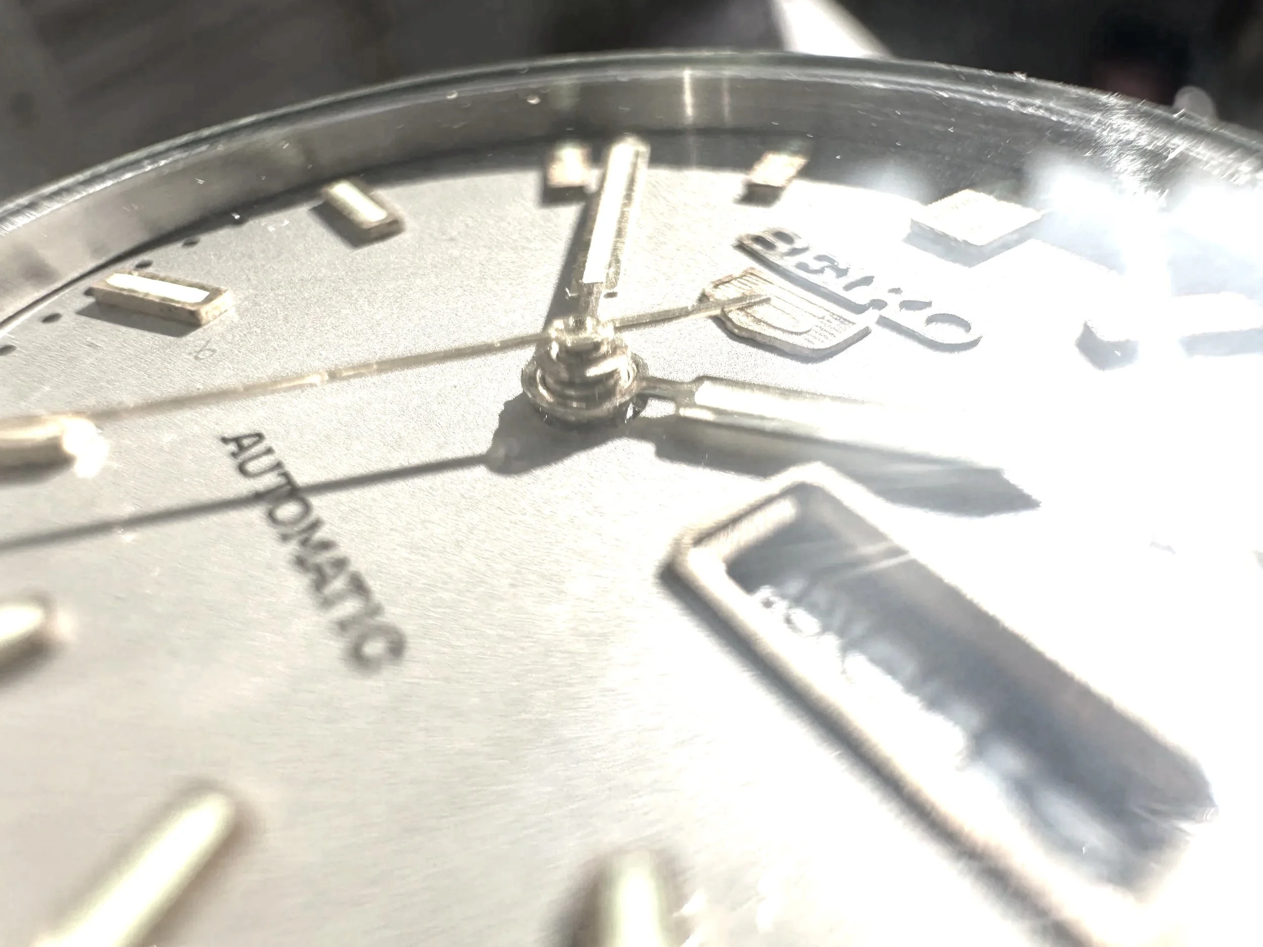 Close-up photo of a watch face showing the time 10 to 2, with visible hour markers, minute and second hands, and part of the watch brand logo.