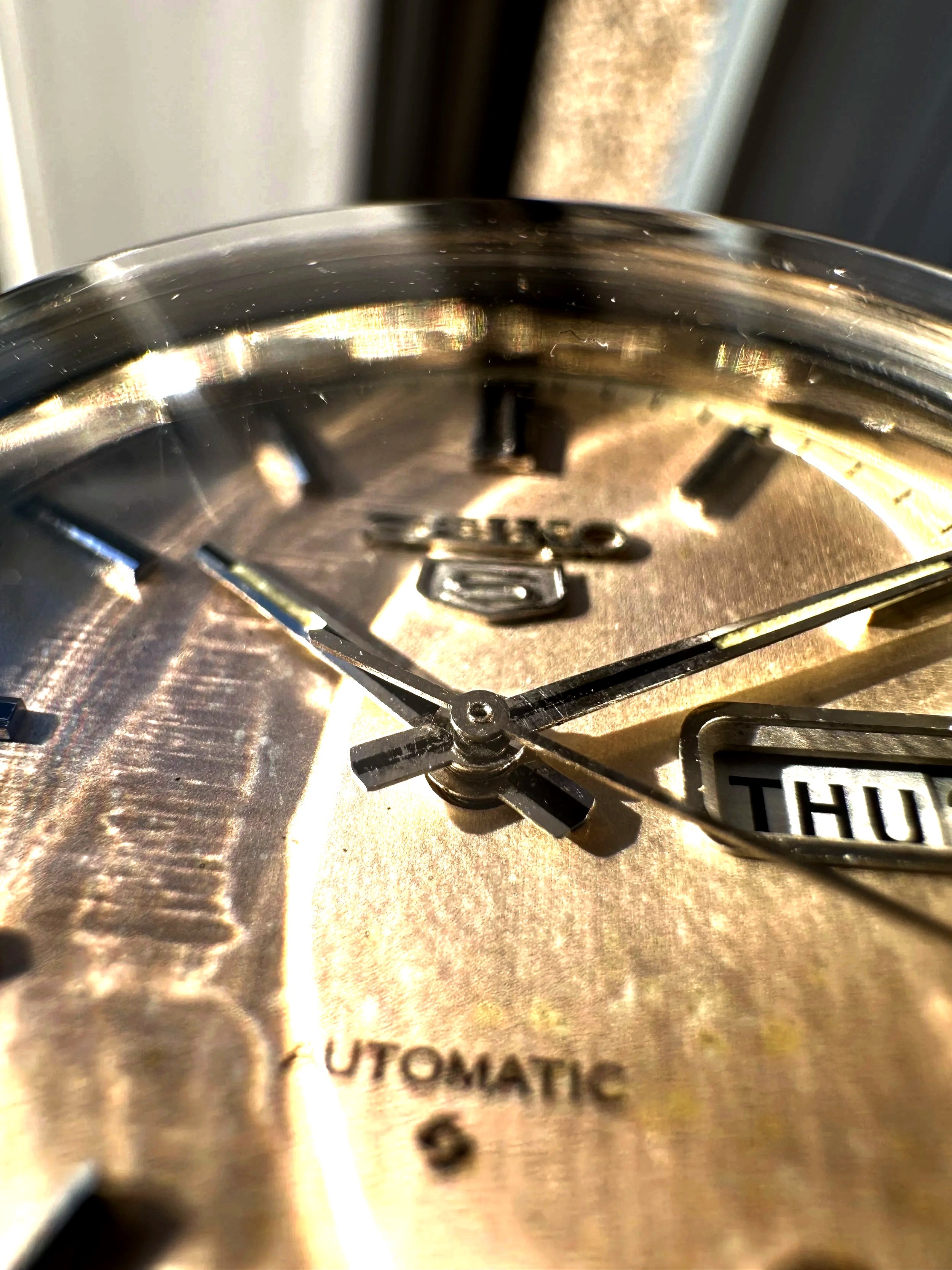 Close-up of a stainless steel clock face, showing black hour and minute hands pointing to approximately 12:03, with the word 'AUTOMATIC' partially visible at the bottom.
