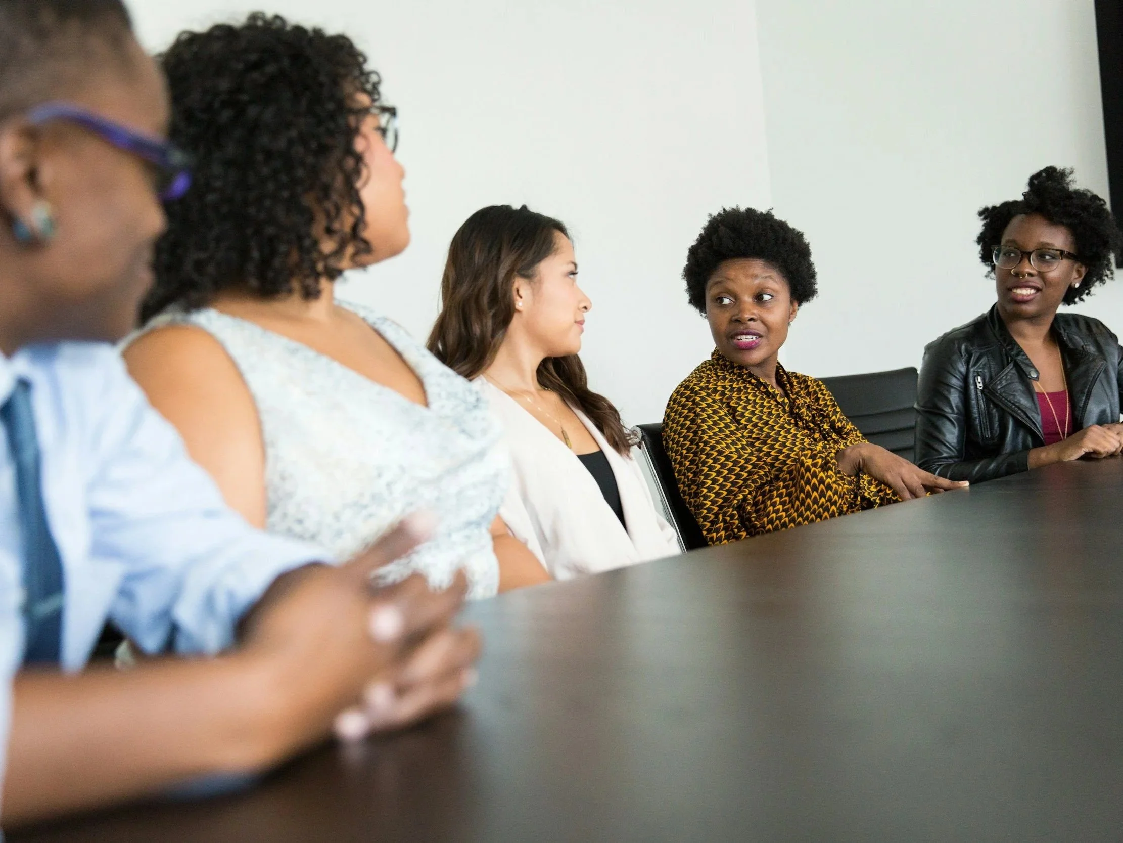 A diverse group of women sitting at a conference table engaged in conversation, with a plain white wall in the background.