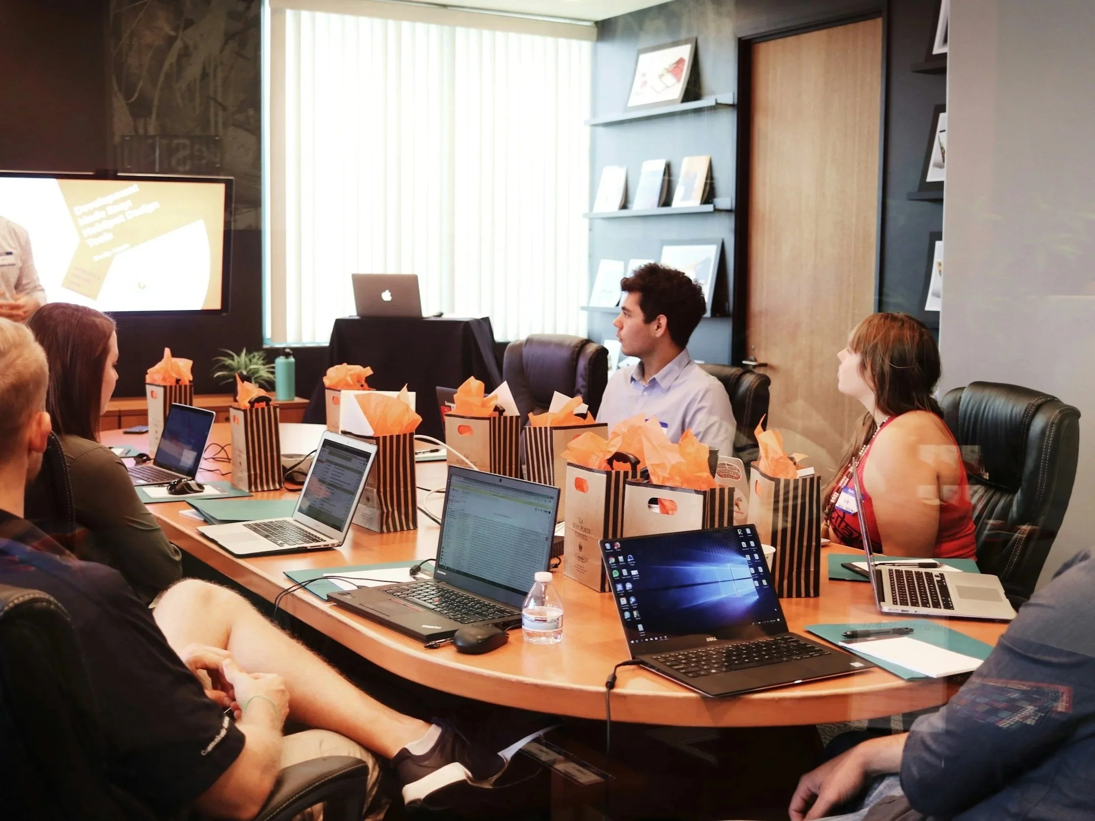 People sitting around a conference table with open laptops, gift bags with orange tissue paper, a bottle of water, and notebooks, during a meeting or presentation in a conference room.