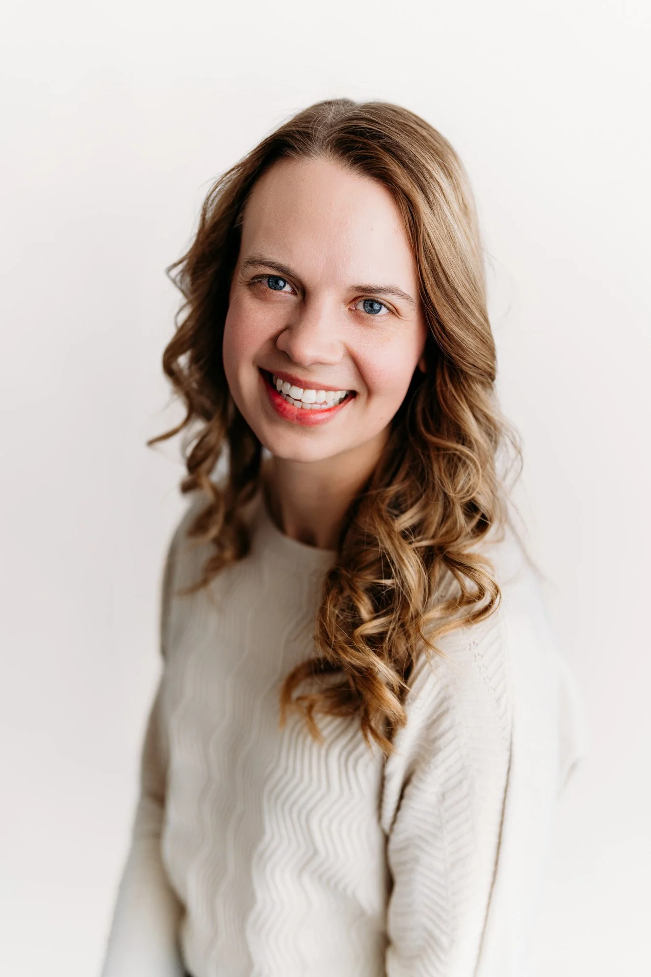 A young woman with blue eyes, light skin, and long wavy brown hair smiling, wearing a cream-colored sweater, against a plain white background.