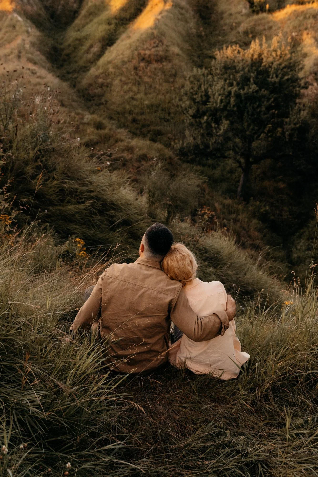 A couple sitting close together in a grassy field near hills, facing away from the camera, during sunset or dusk.